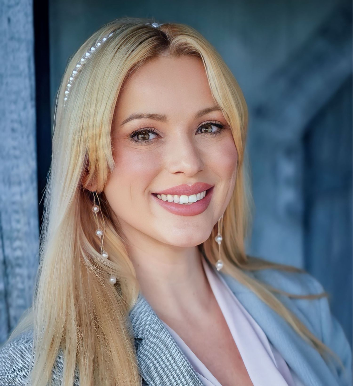 Beautiful blonde woman with pearl headband and earrings smiles warmly in close-up portrait photo.