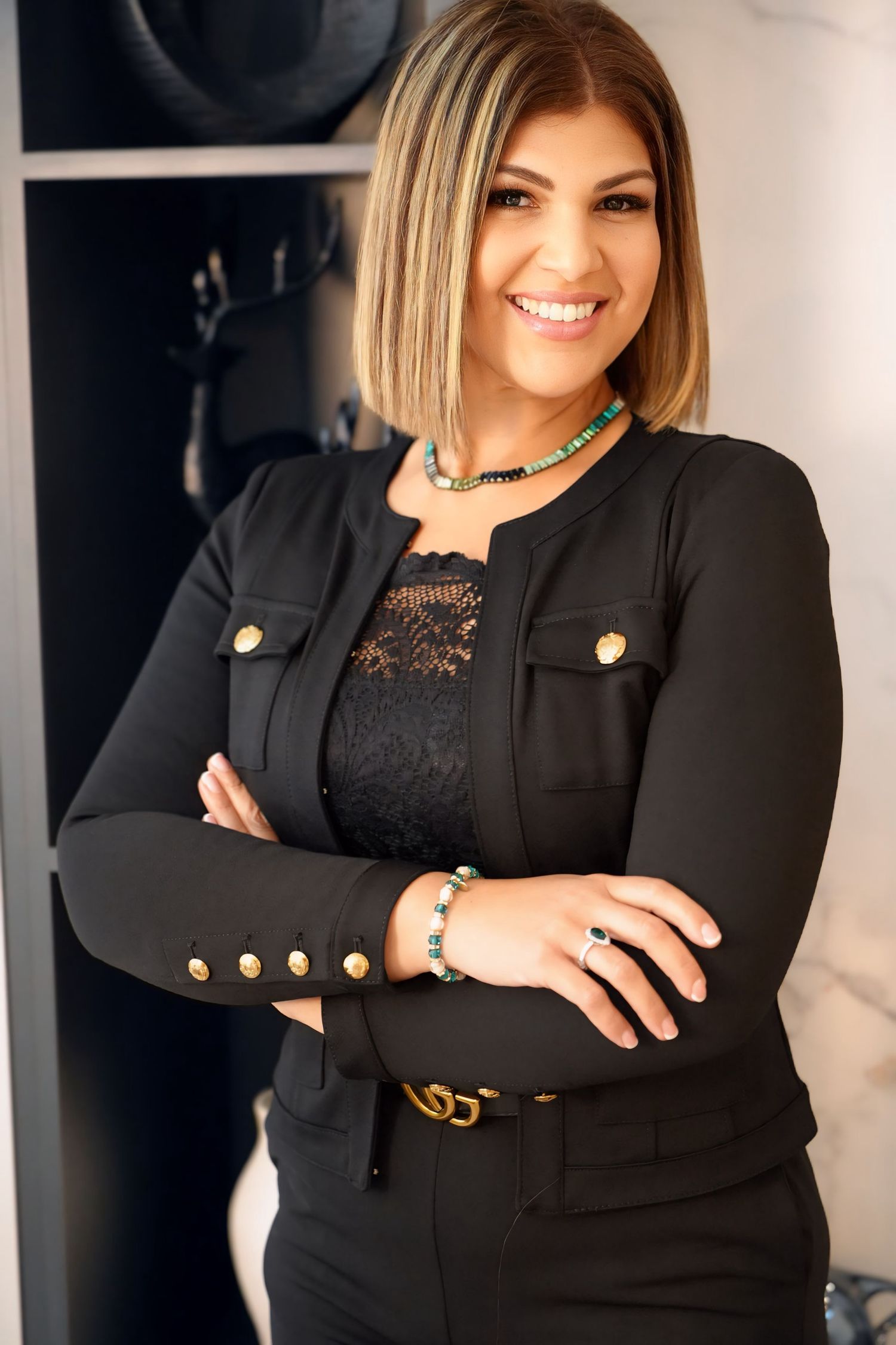 Smiling woman in black blazer with gold buttons poses with arms crossed in bright interior.