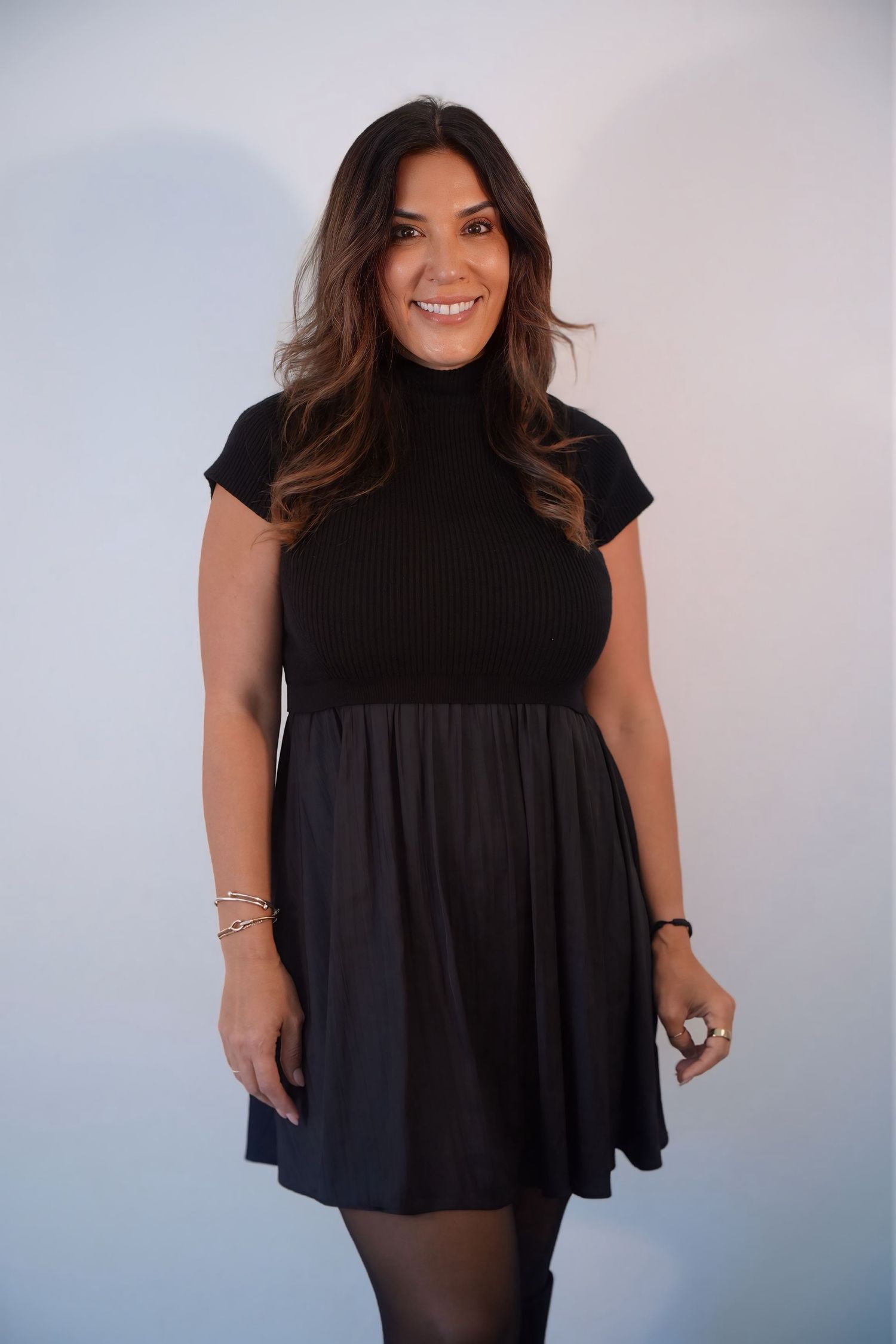 Smiling brunette woman in black turtleneck mini dress posing against light gray background.