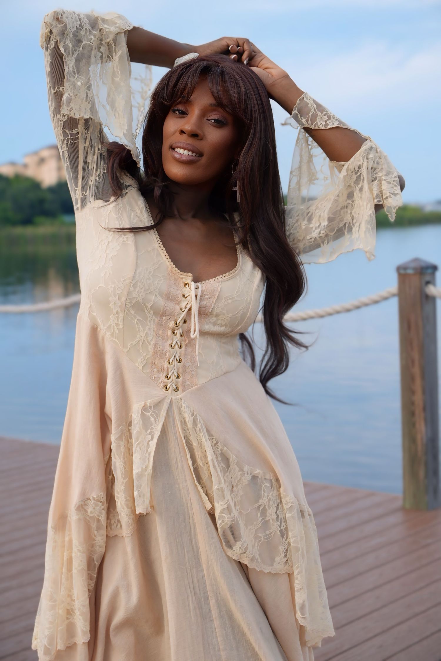 Elegant woman in cream lace boho dress poses on waterfront dock at dusk.
