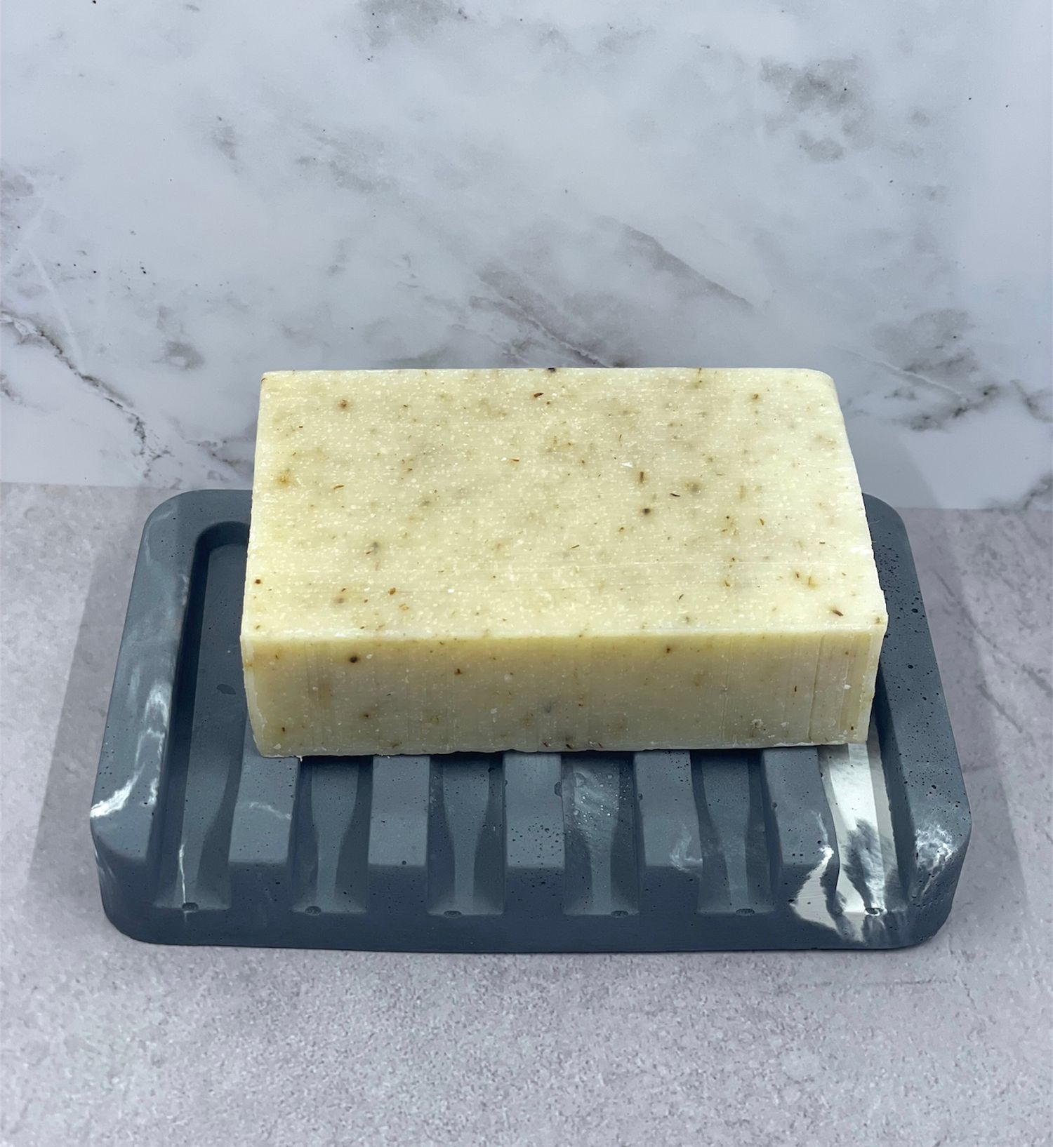 Handmade natural herb bar soap resting on gray ribbed ceramic soap dish on marble surface.