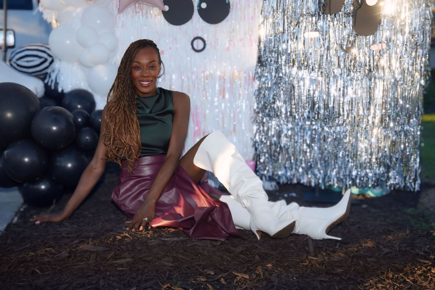Woman in pink skirt and white boots sitting on ground at festive party with black and silver balloon decorations.