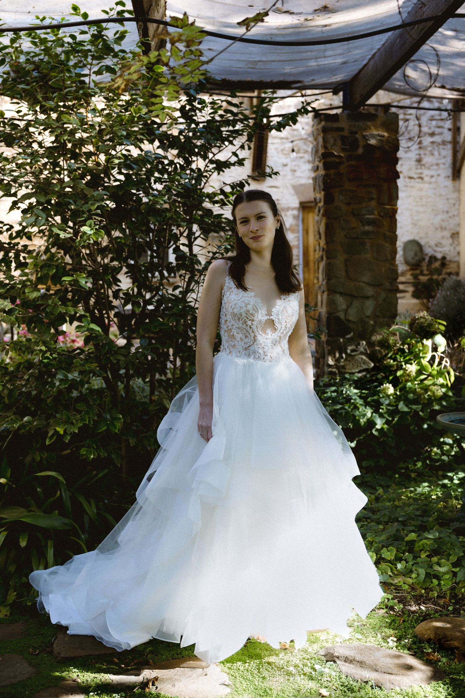 Bride in elegant white layered tulle ball gown standing in lush garden with stone wall backdrop.