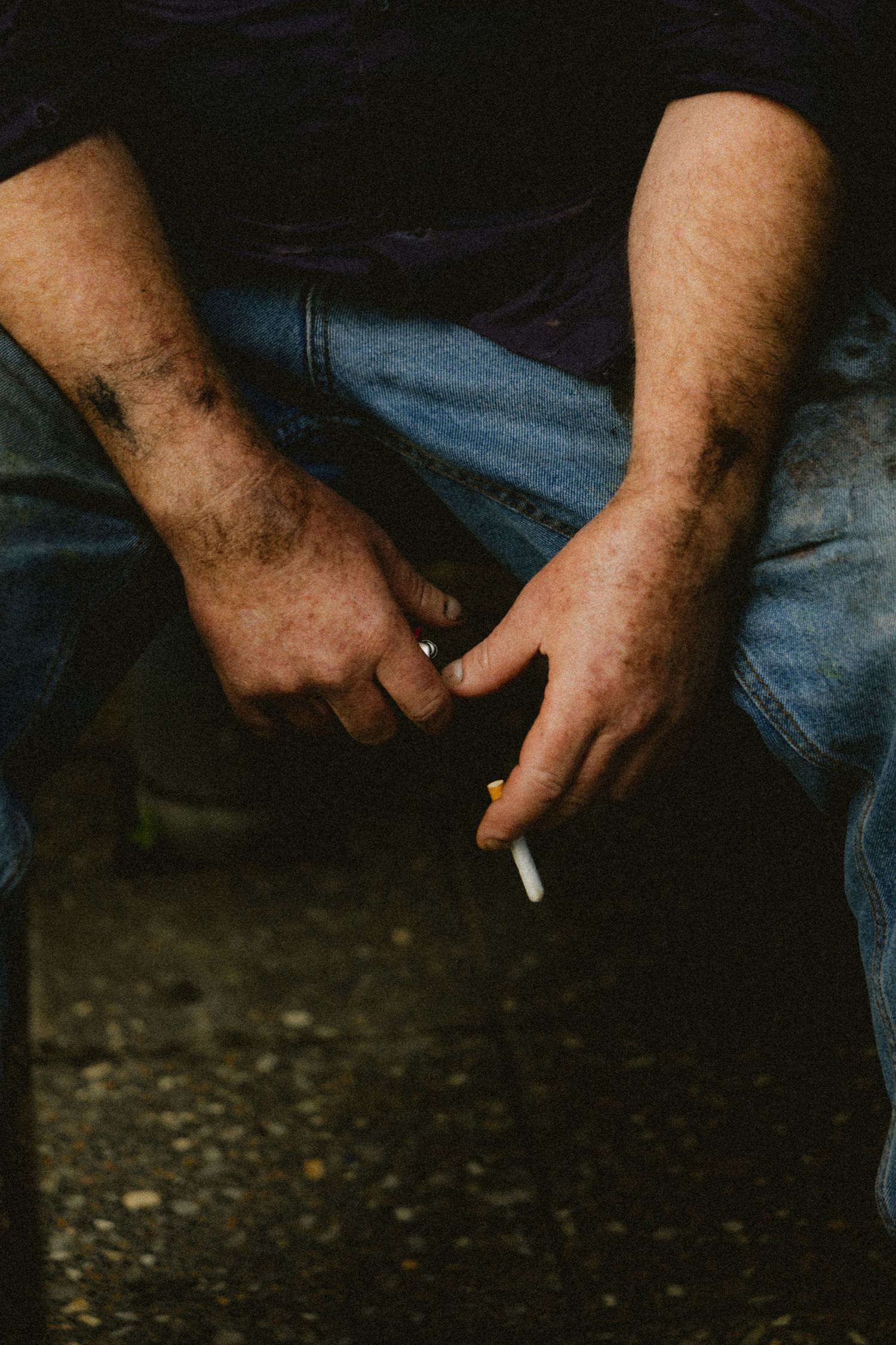 Close-up of dirty male hands holding a cigarette resting on denim jeans.