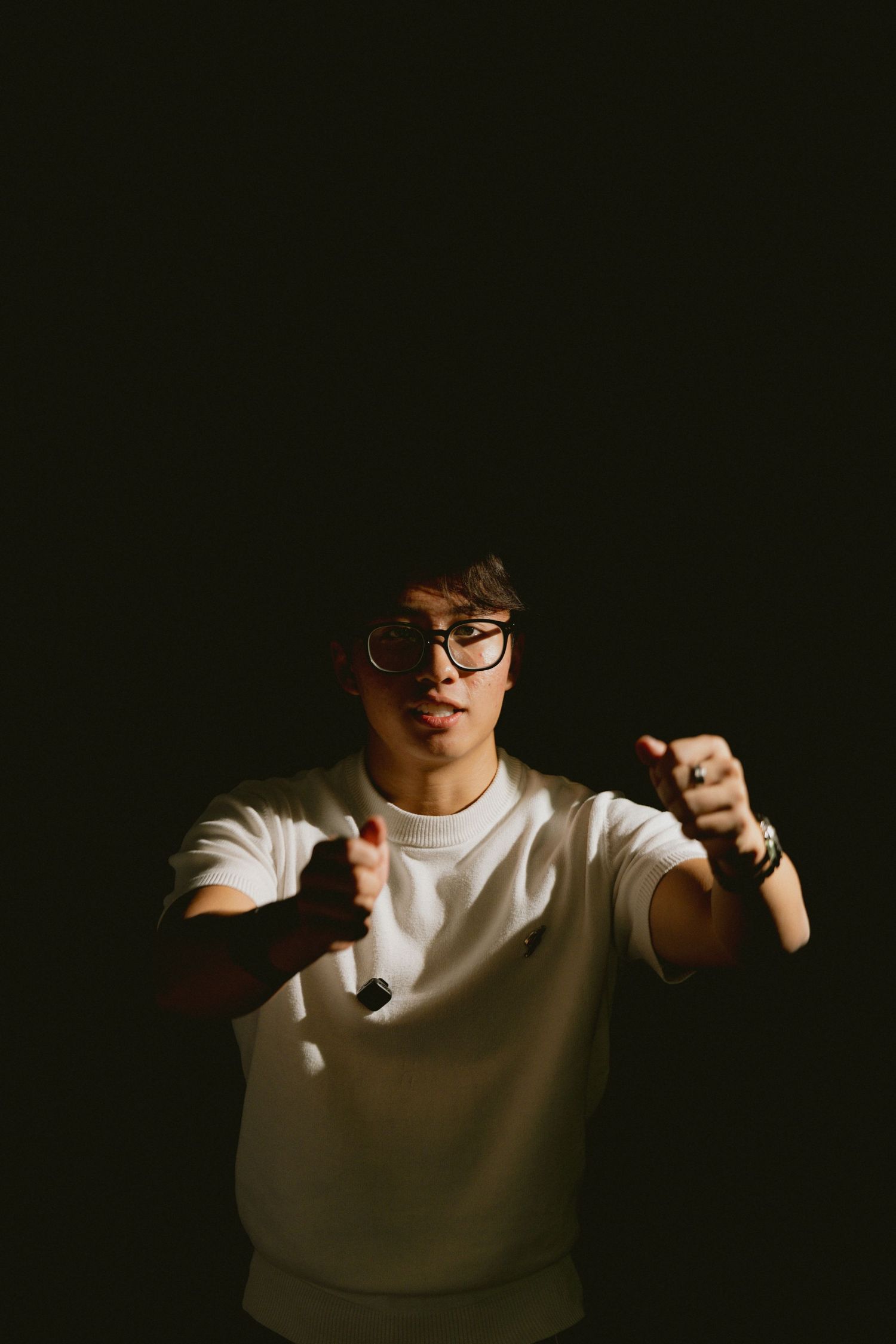 Young man in white t-shirt and glasses posing with fists raised against dark background.