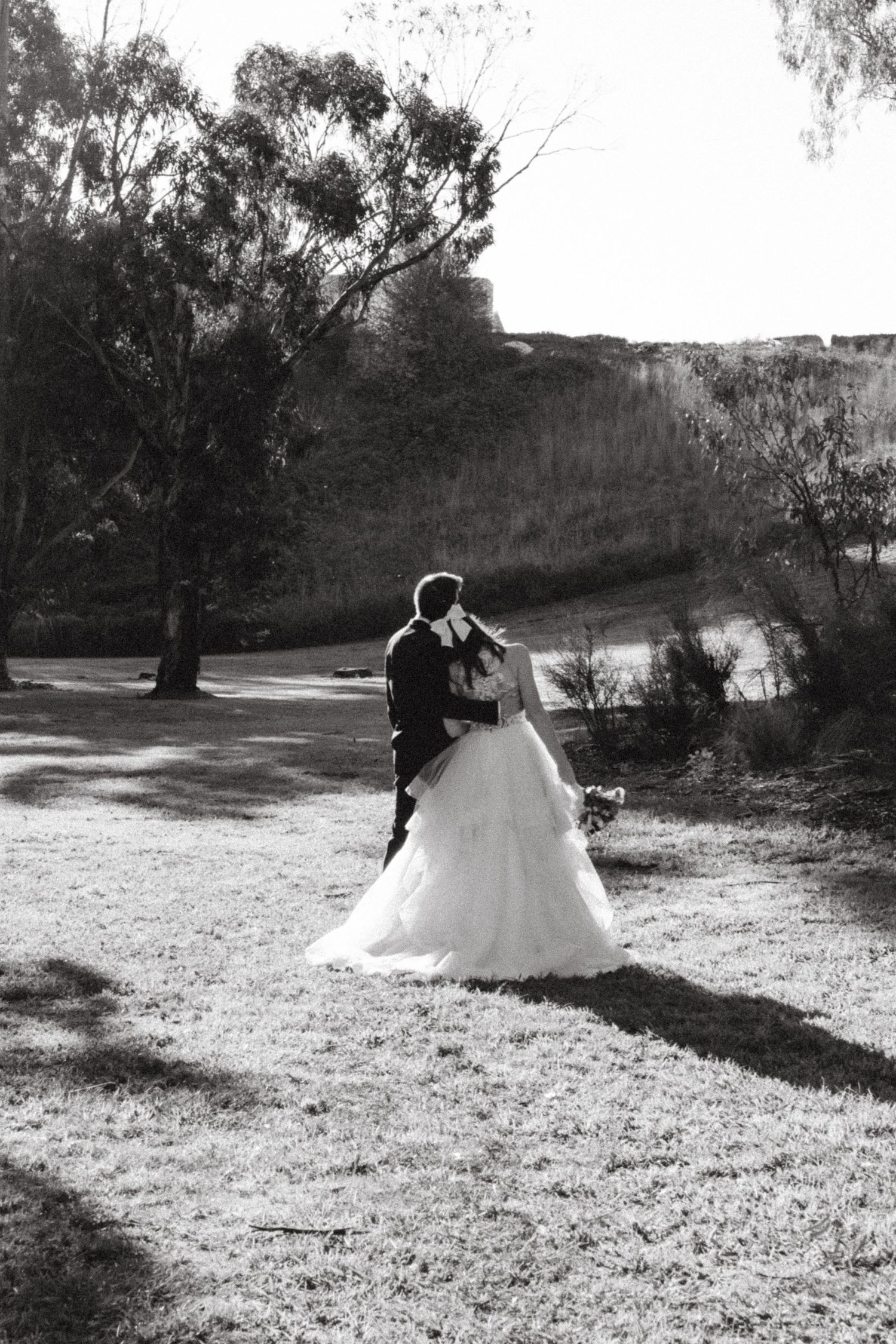 Black and white photo of bride and groom embracing in an open field with trees.