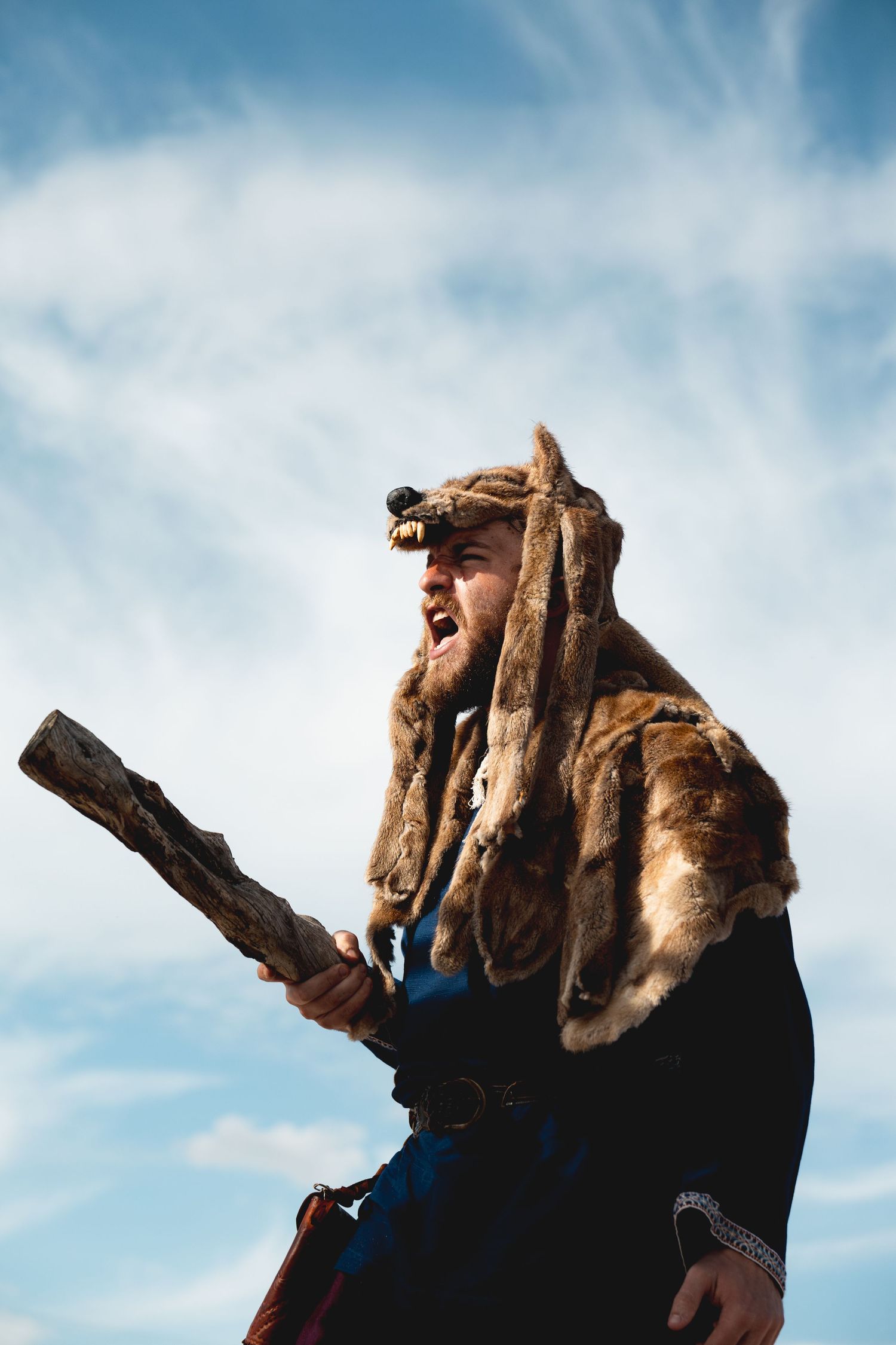 Bearded man in fur Viking costume yells while holding wooden club against cloudy sky.