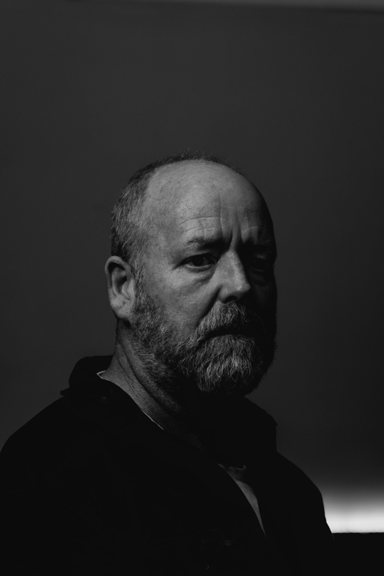 Black and white portrait of a bearded bald man with a contemplative expression in moody lighting.