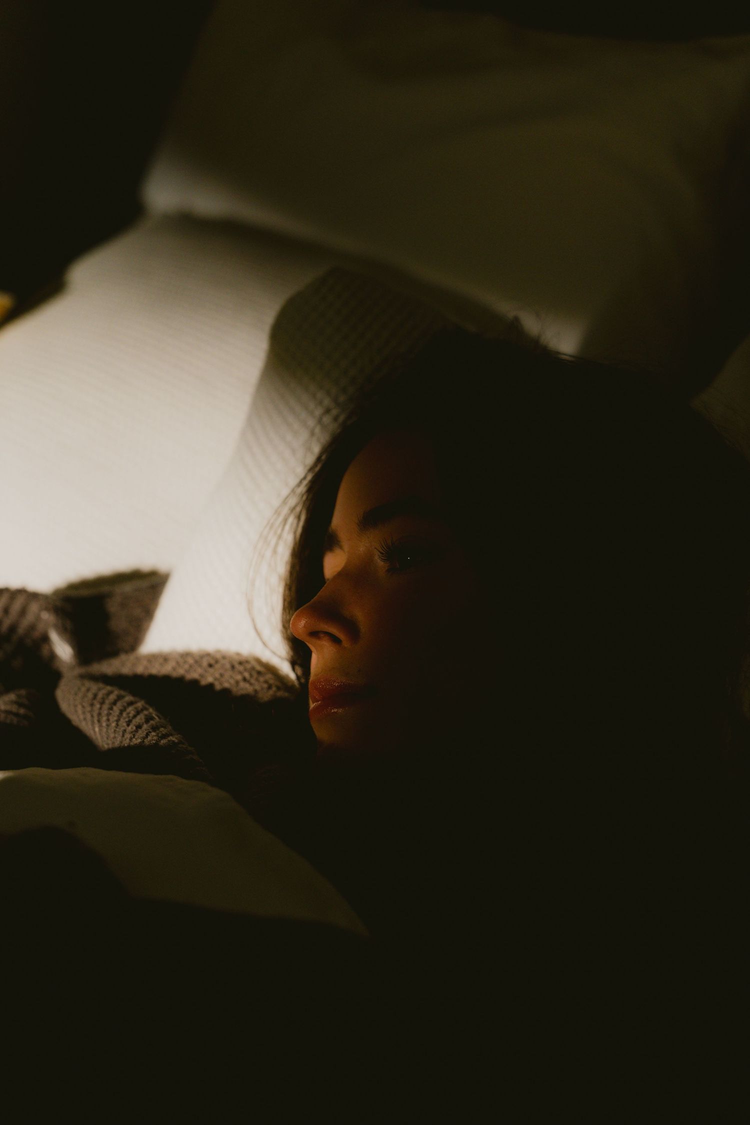 Woman with dark hair lying in bed, partially illuminated by warm light in a dark room.