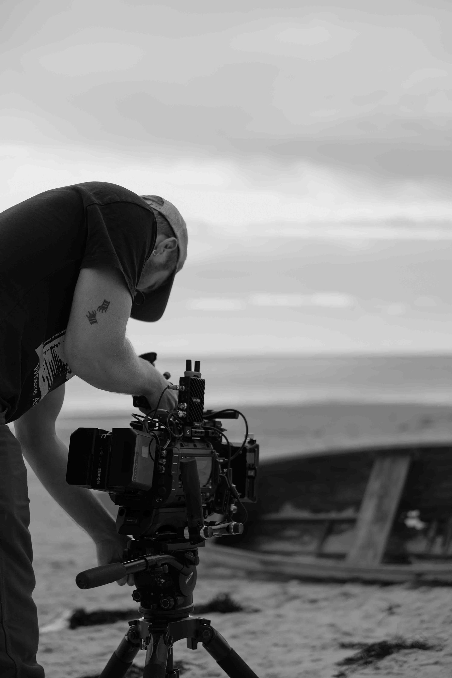 Cameraman operating a professional film camera on a tripod at a beach during overcast conditions.