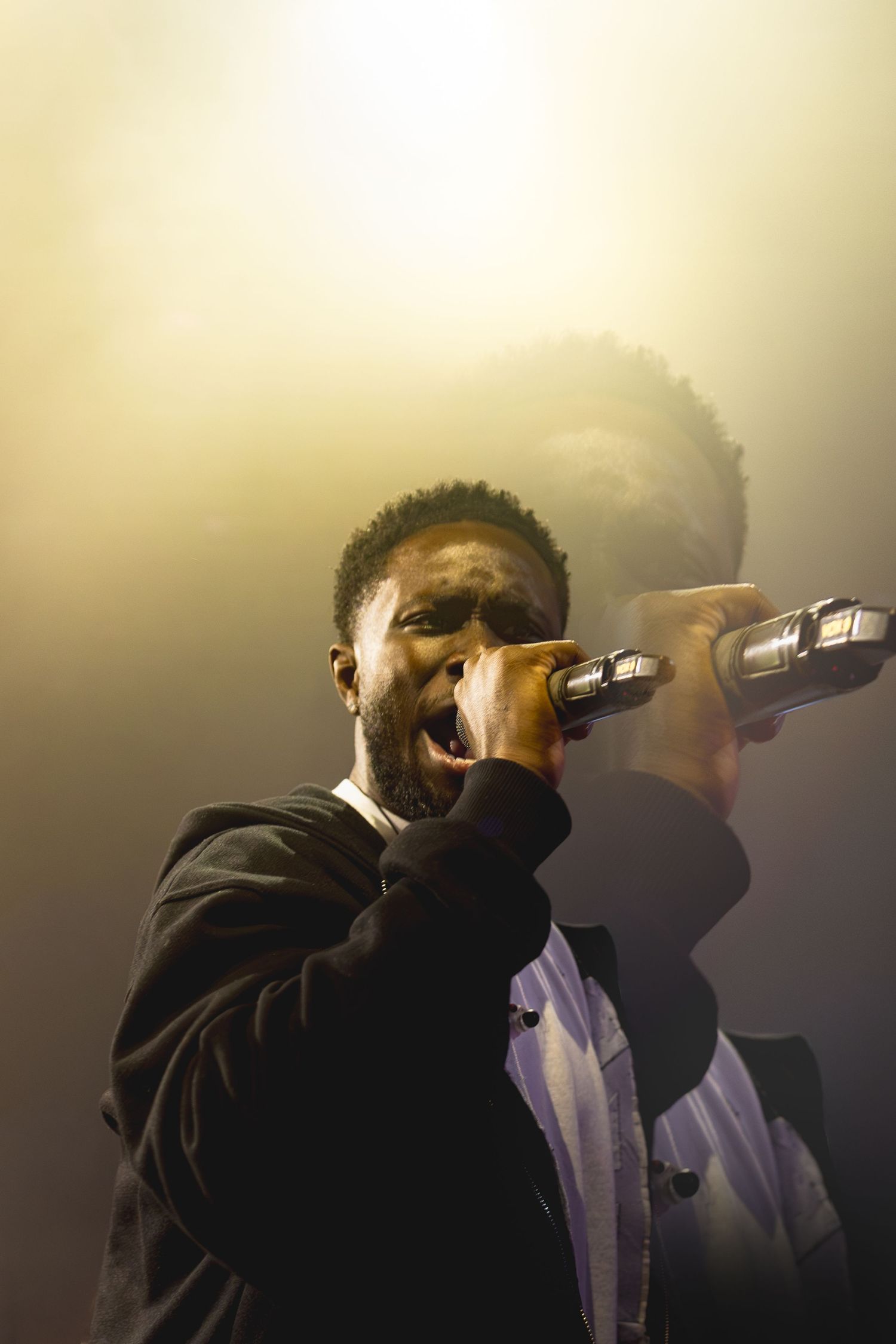 A Black male singer performs passionately on stage, holding a microphone under dramatic yellow stage lighting.