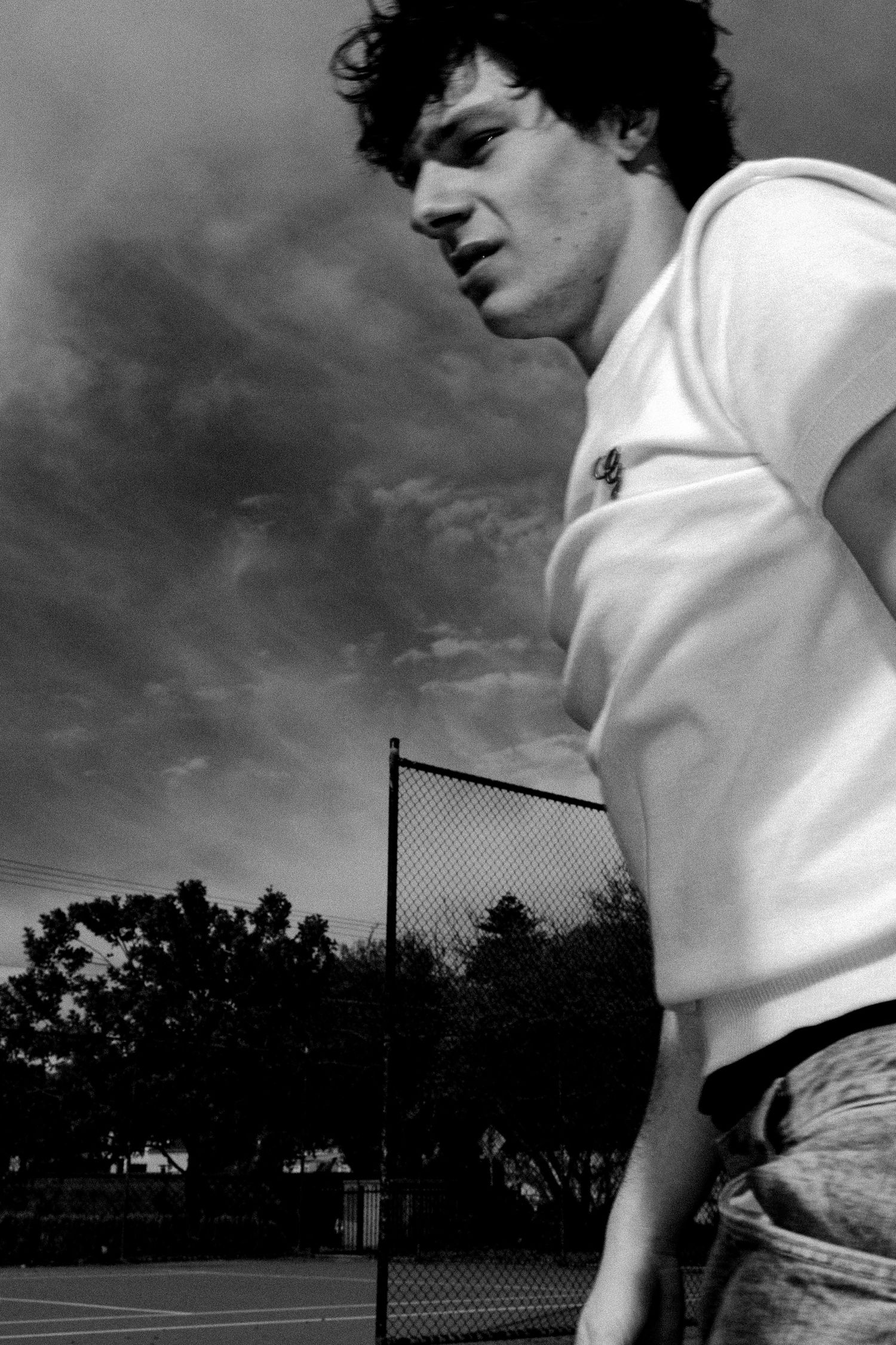 Black and white photo of a young man in a white t-shirt on a tennis court with dramatic cloudy sky.