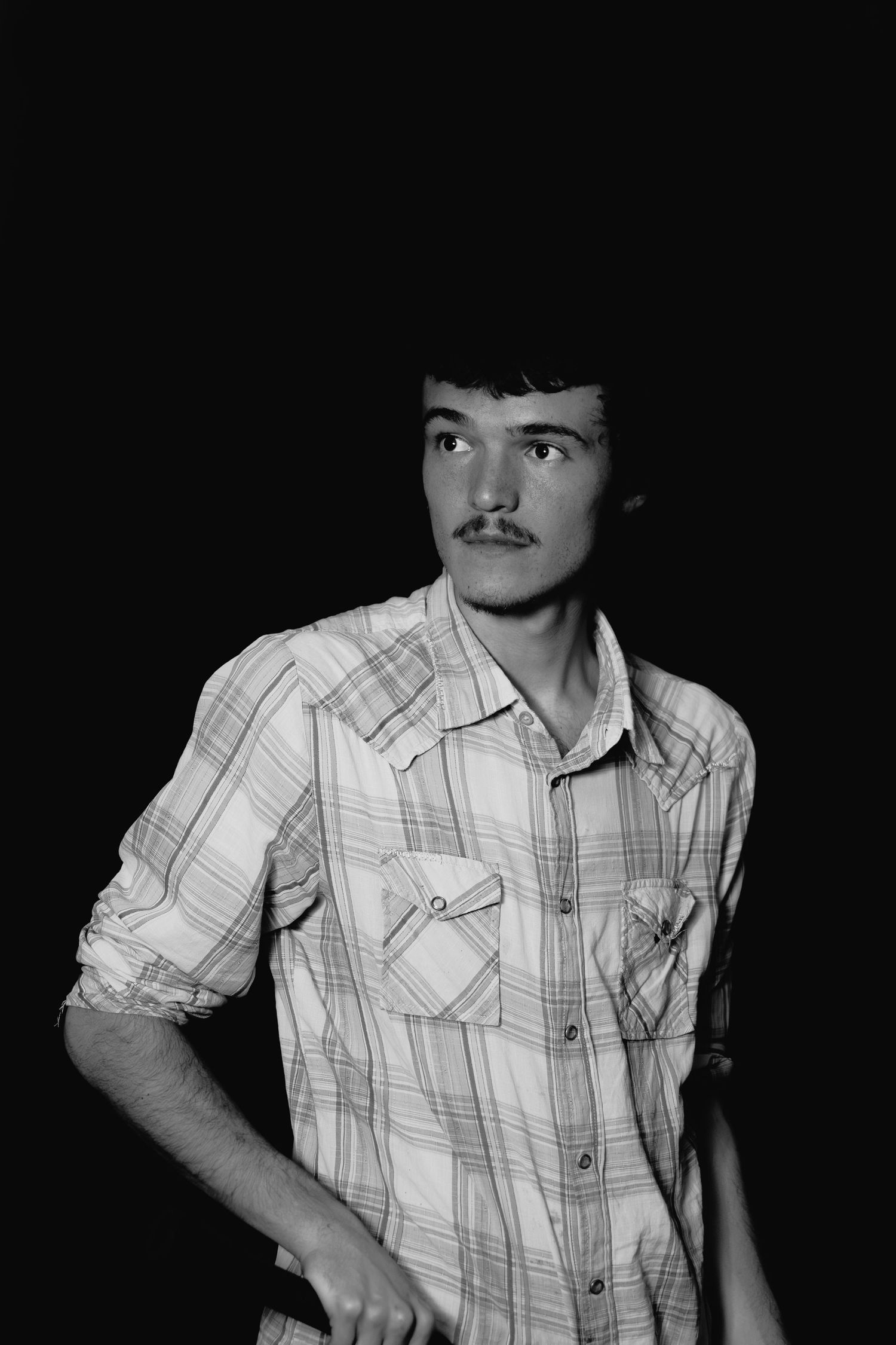Black and white portrait of a young man with mustache wearing a plaid shirt against a dark background.