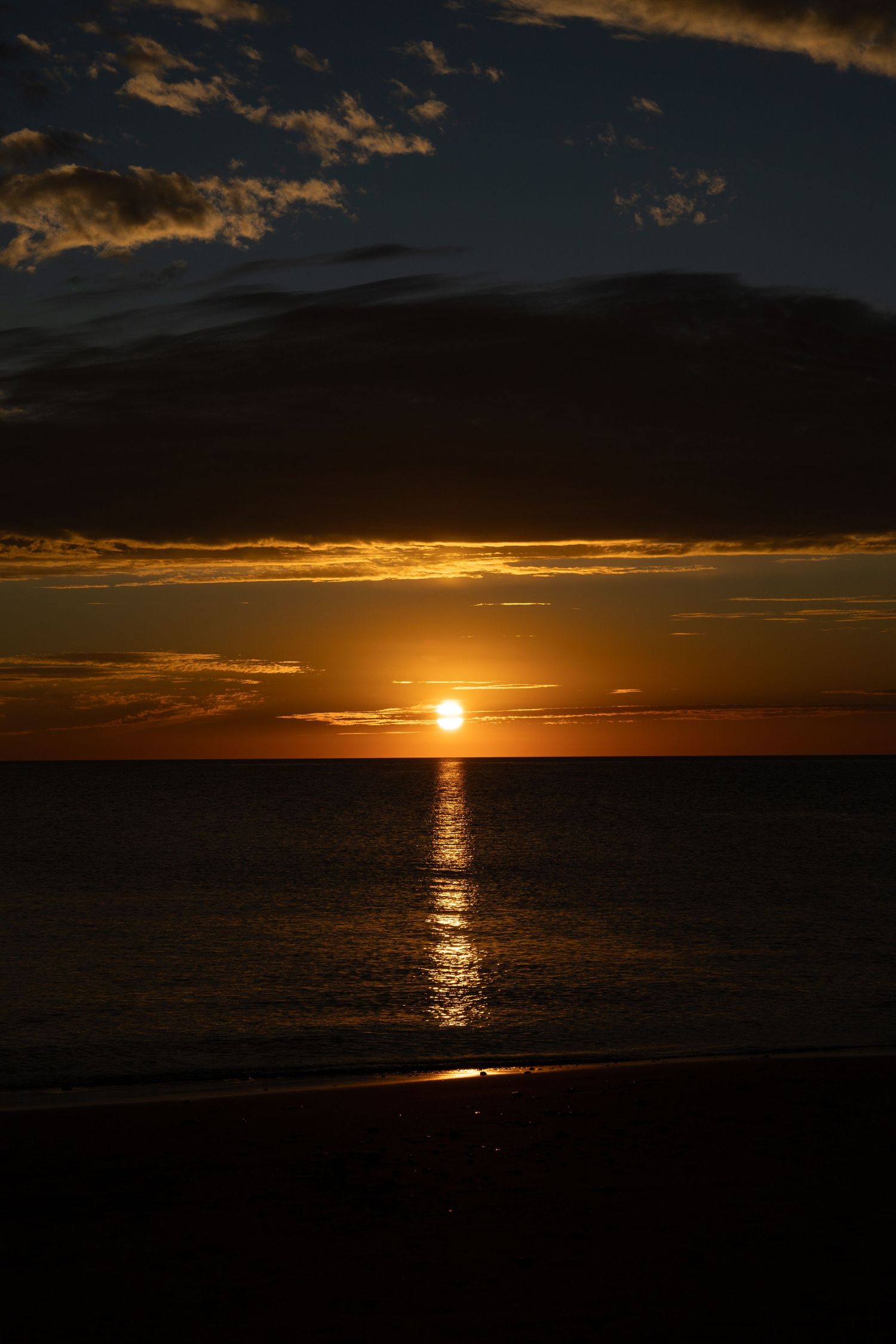 Dramatic golden sunset over calm ocean with dark clouds and sun reflecting on water surface.