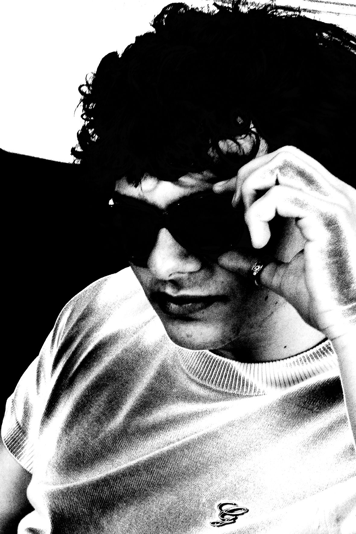 Black and white portrait of a young man with curly hair adjusting sunglasses, wearing a white sweater.