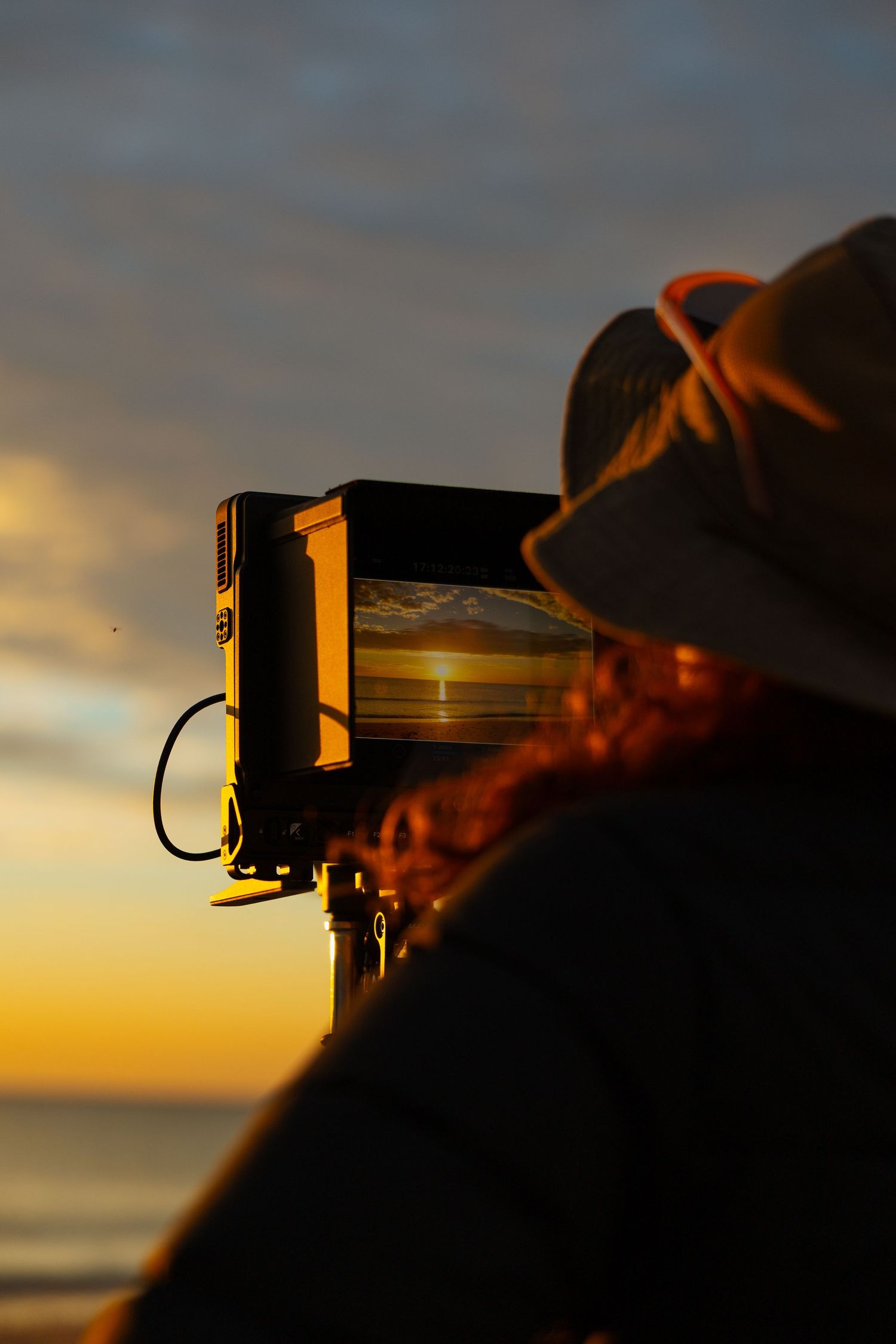 Silhouette of a person wearing a hat watching a stunning ocean sunset reflected on a camera monitor.