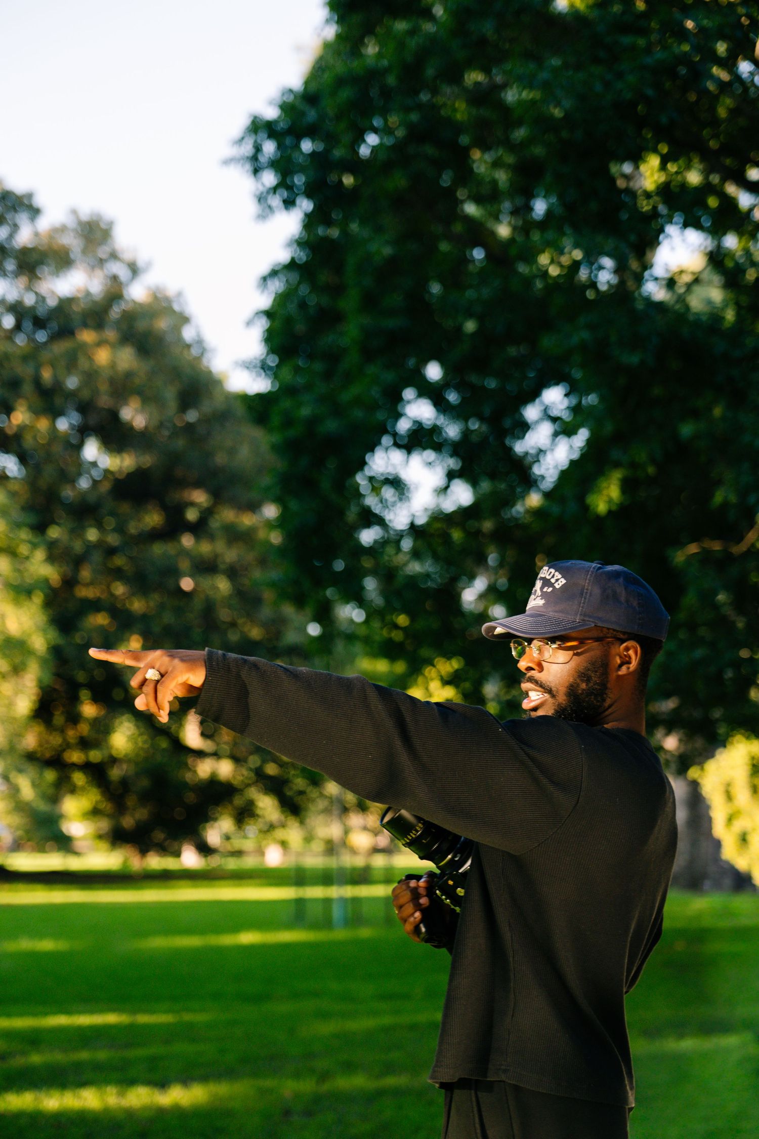 Man in black outfit and cap pointing into the distance while holding a camera in a park.