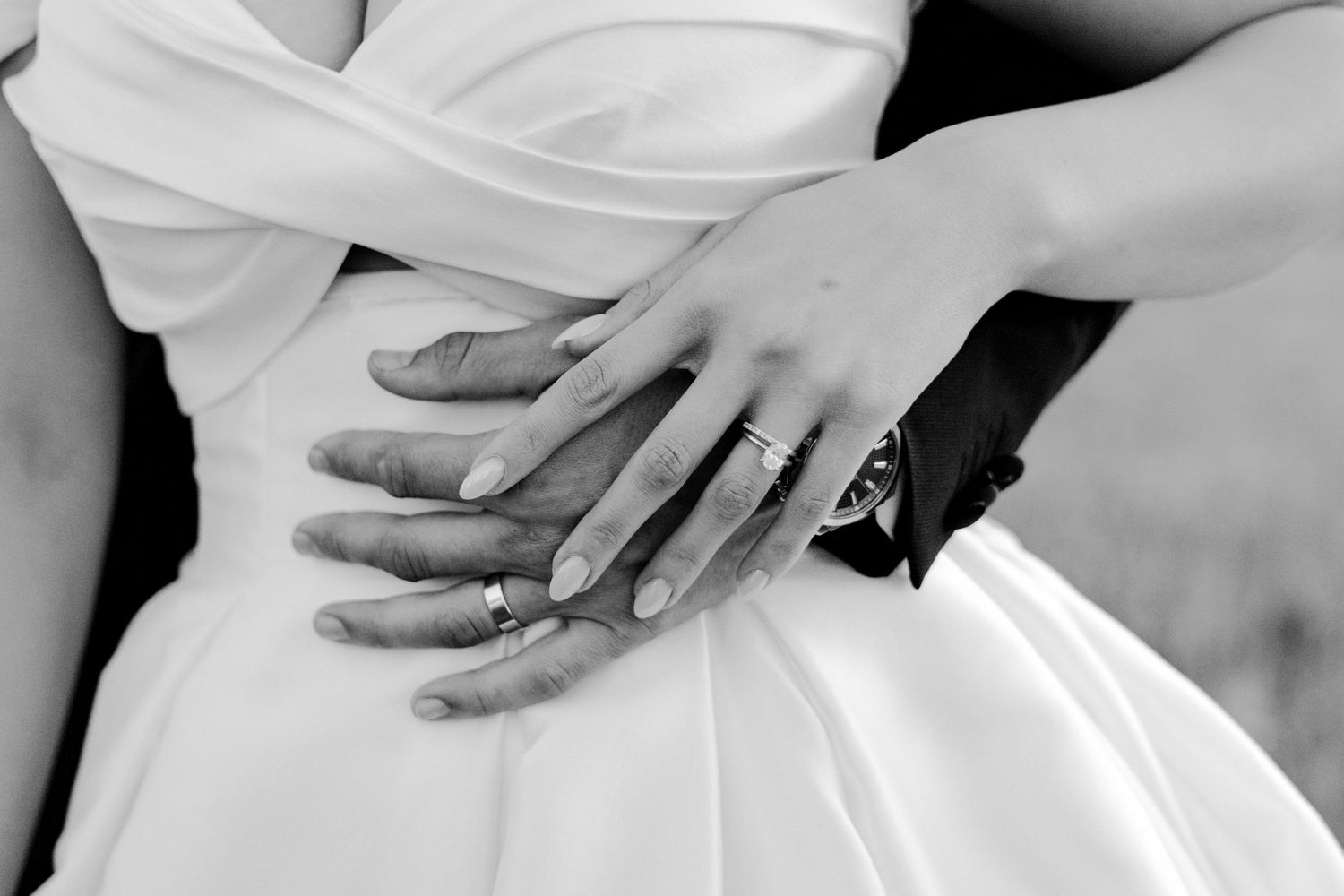 Black and white photo of newlyweds hands with wedding rings embracing at the waist in a wedding dress.