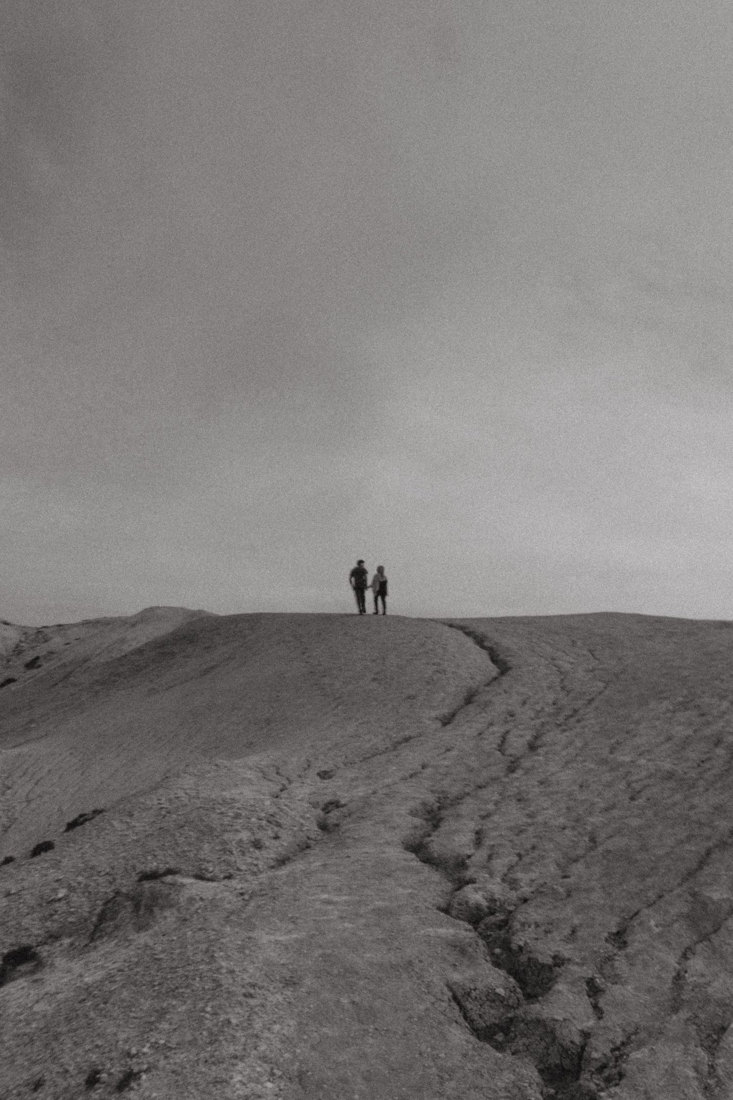 Two silhouetted figures stand atop a rocky ridge under an overcast sky in this black and white landscape photo.