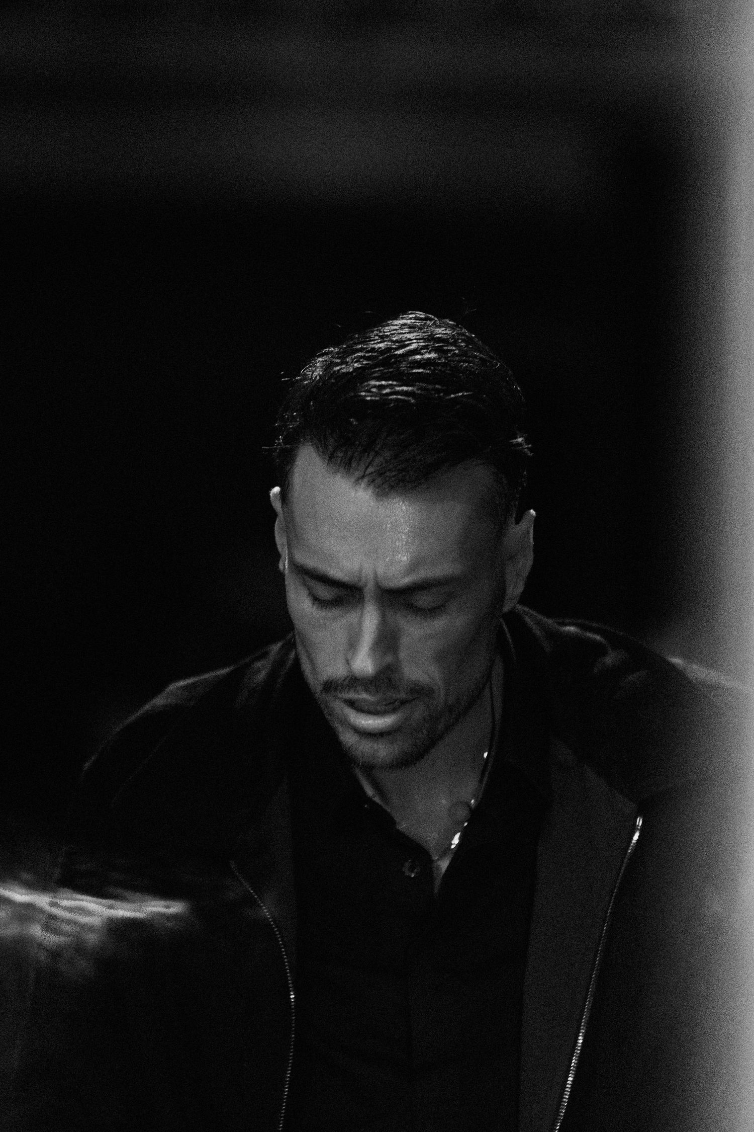 Black and white portrait of a man with slicked-back hair and stubble, looking downward in dramatic low-key lighting.