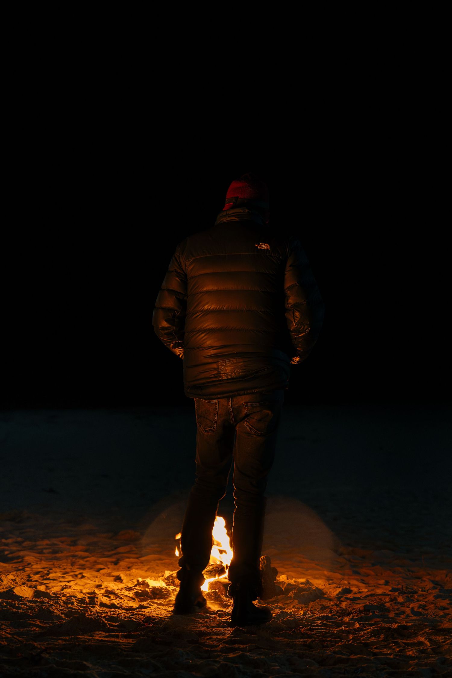 Person in North Face jacket and red beanie standing before a small fire on dark snowy ground.