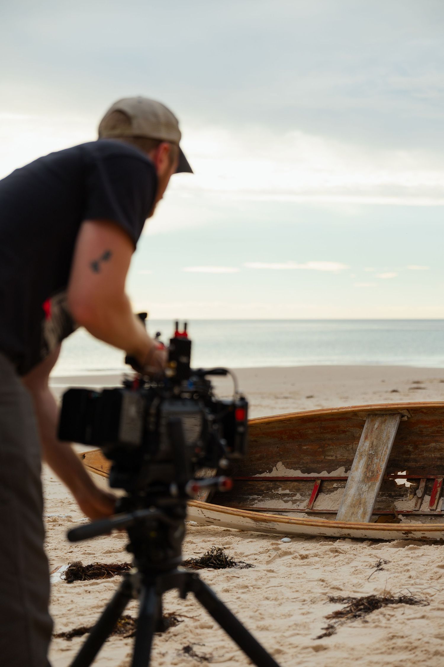 A cameraman frames a shot of an old wooden boat on a beach at dusk.