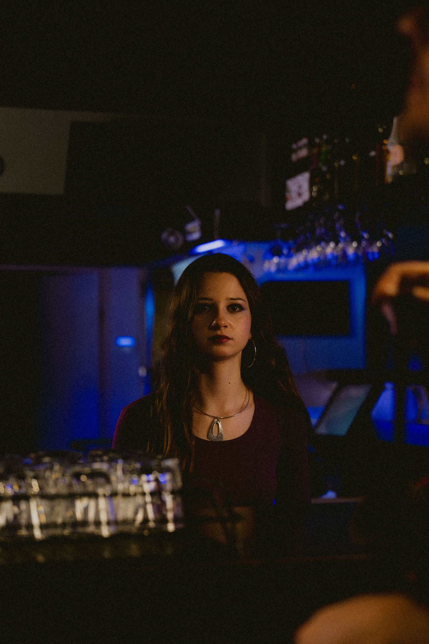 Young woman in dark red top standing at a bar counter under moody blue lighting.