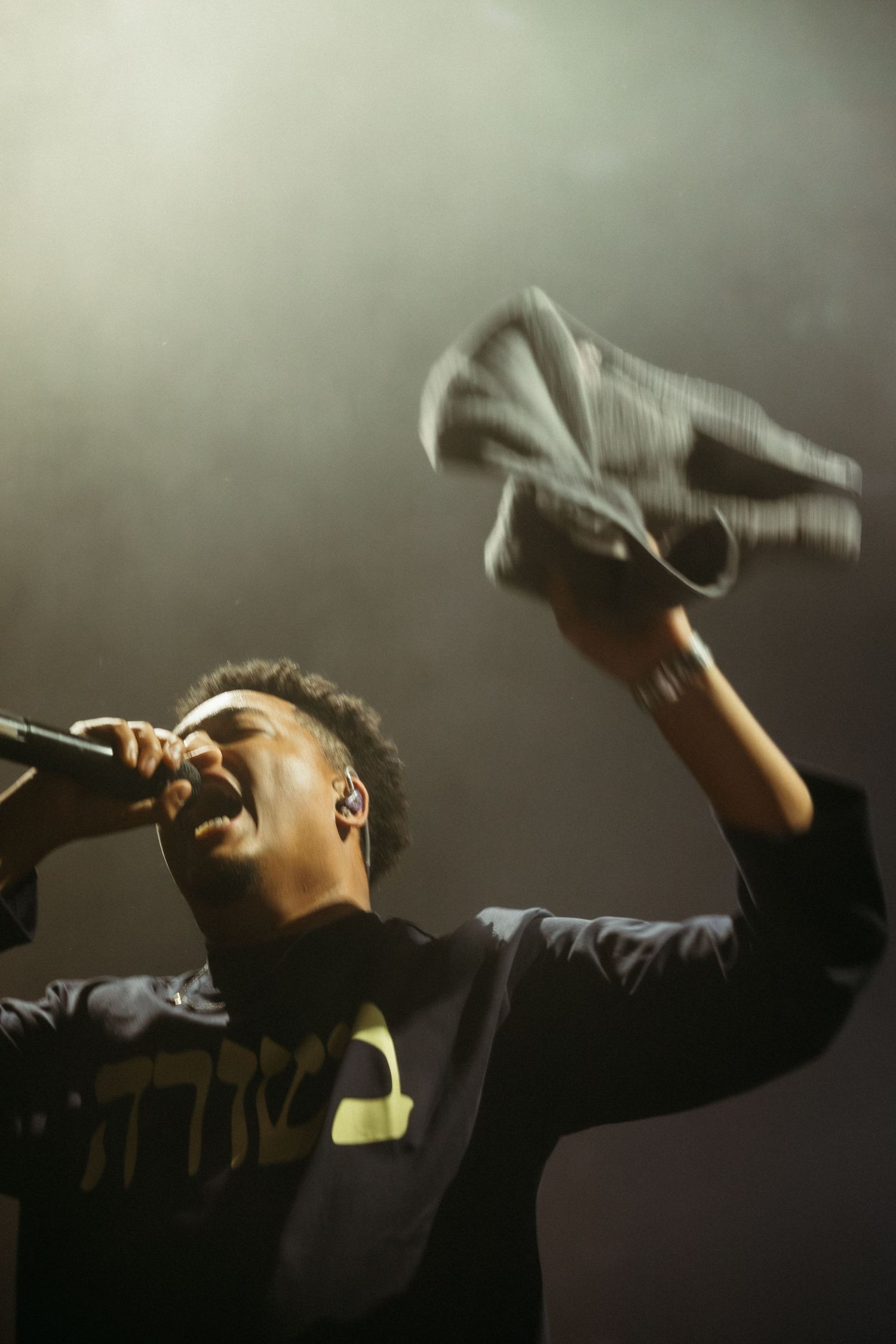 Young male performer singing passionately on stage, holding microphone and waving jacket under dramatic spotlight.