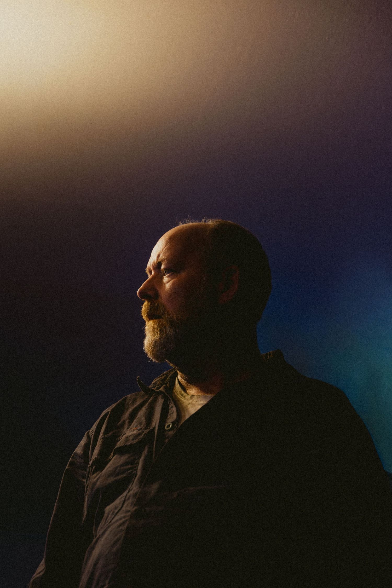 Dramatic side profile of a bearded middle-aged man lit by warm golden light against a dark blue background.