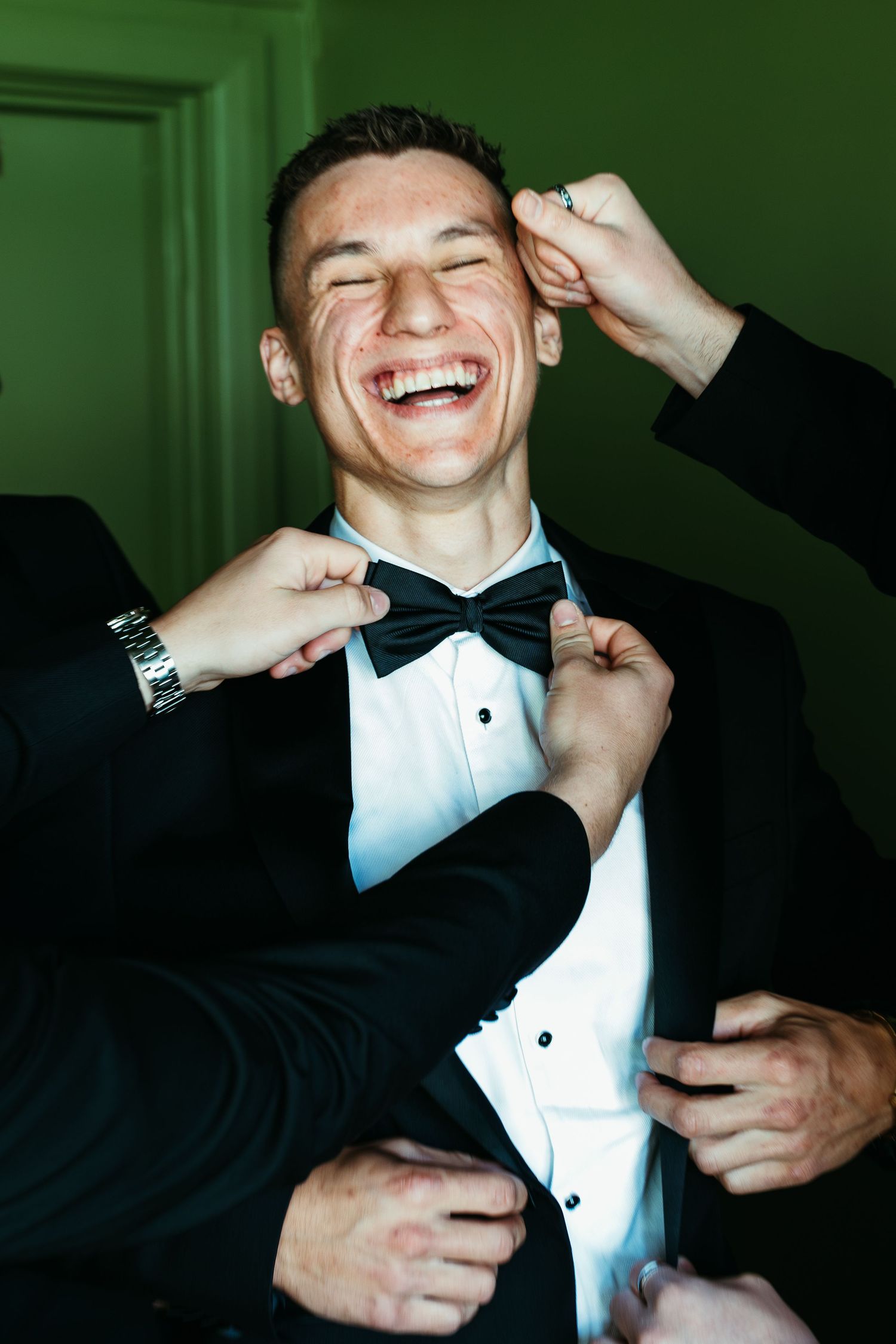 Laughing groom in tuxedo being helped by groomsmen adjusting bow tie and jacket before wedding ceremony.