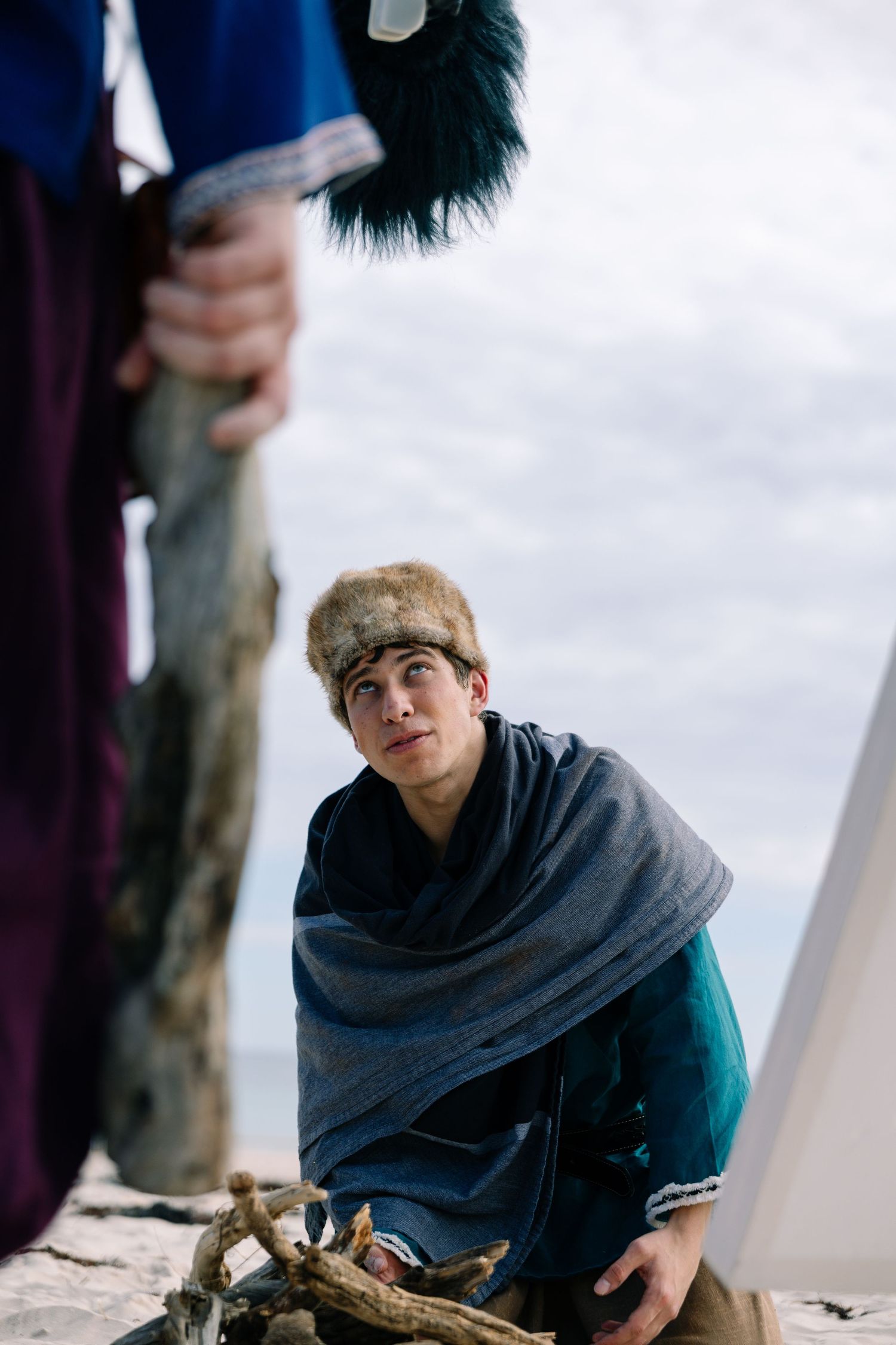 Young man in fur hat and grey cloak kneeling on sandy beach in Viking costume.