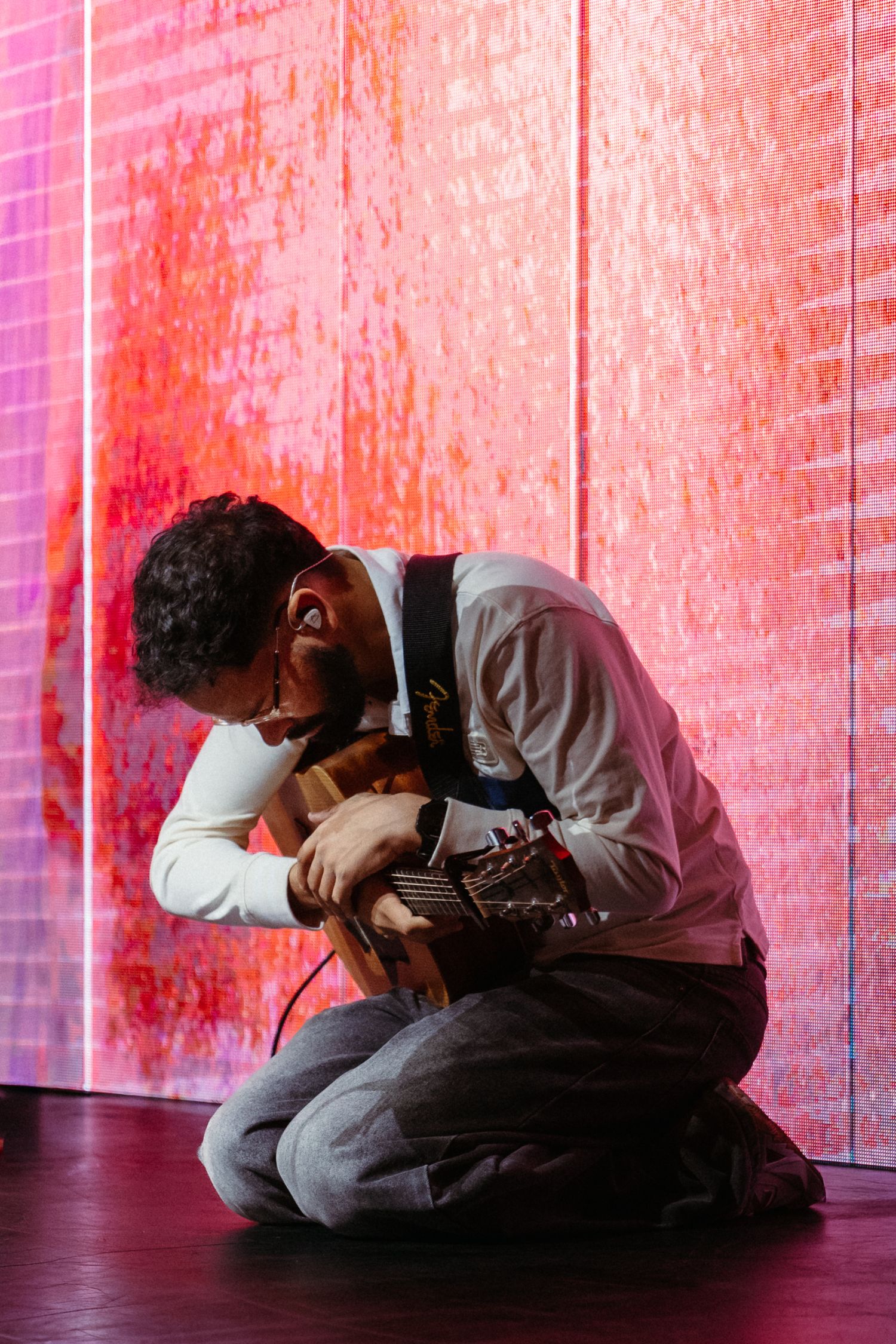 Male guitarist kneels on stage floor holding guitar in a moment of worship under red LED lighting.