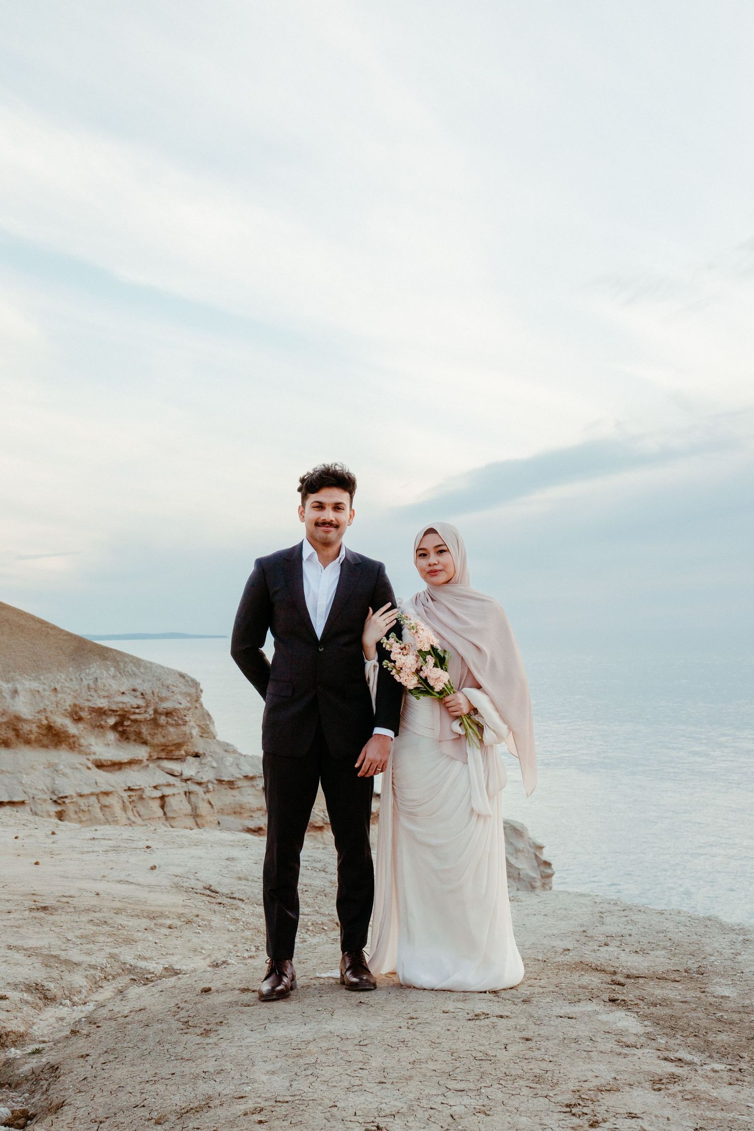 A couple in wedding attire poses on rocky cliffs by the sea, groom in suit, bride in white dress and hijab holding bouquet.