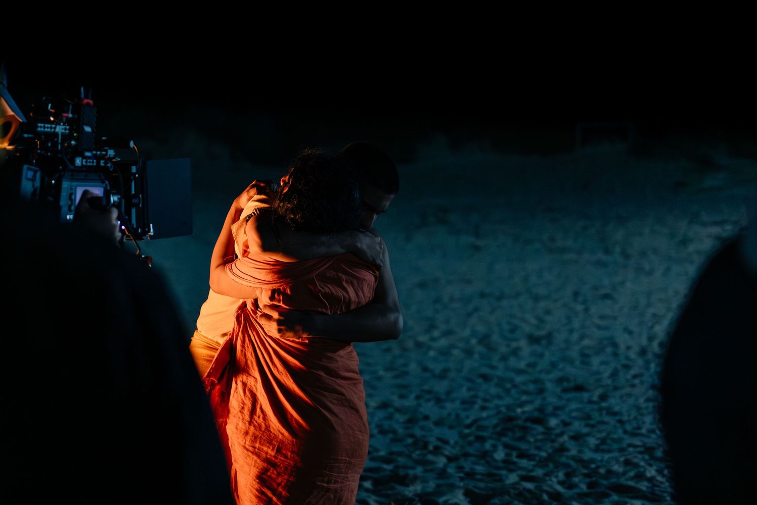 Two people in orange robes share an embrace outdoors at night, lit by warm ambient light.