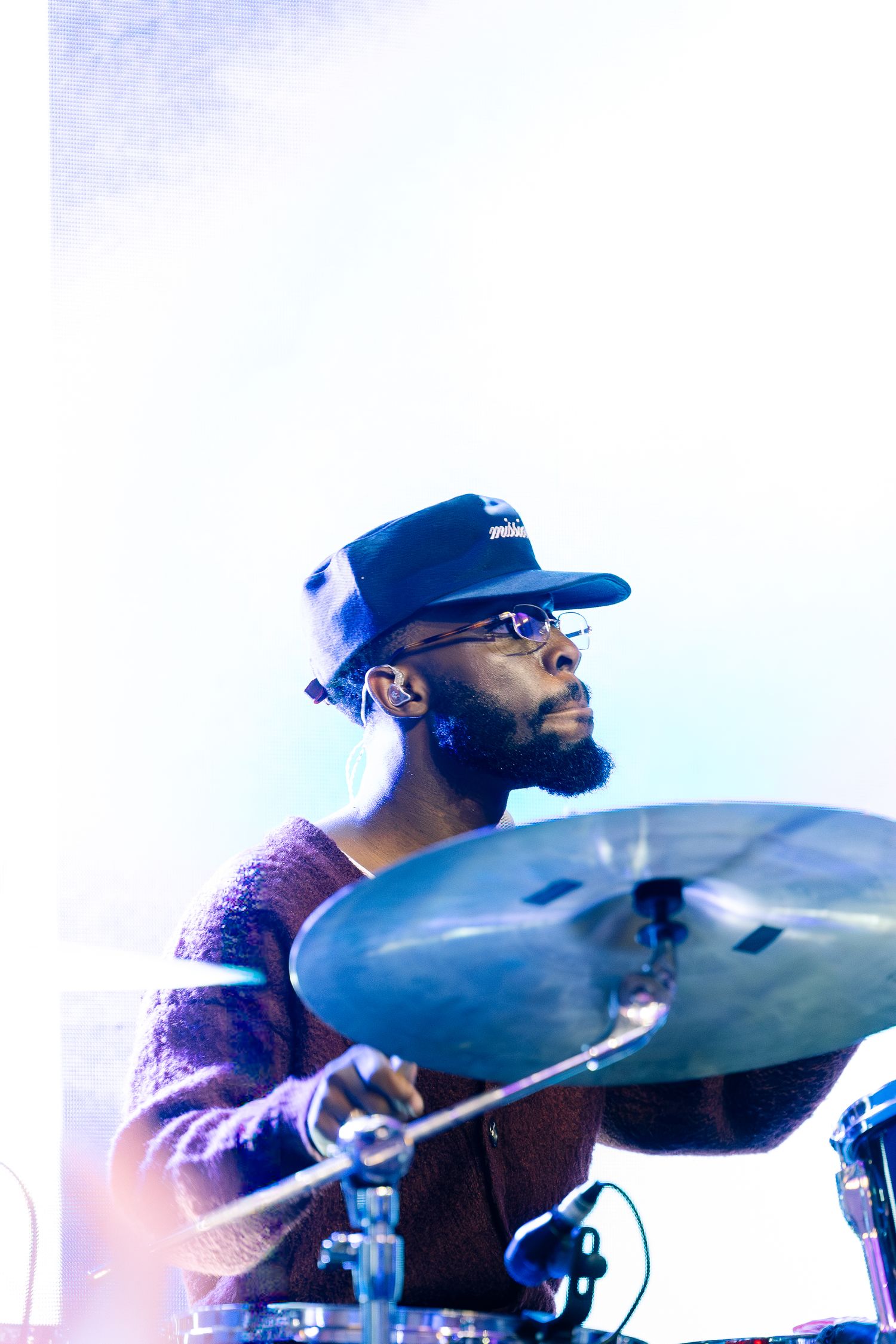 Bearded drummer wearing cap and glasses playing cymbals on stage with bright backlighting.