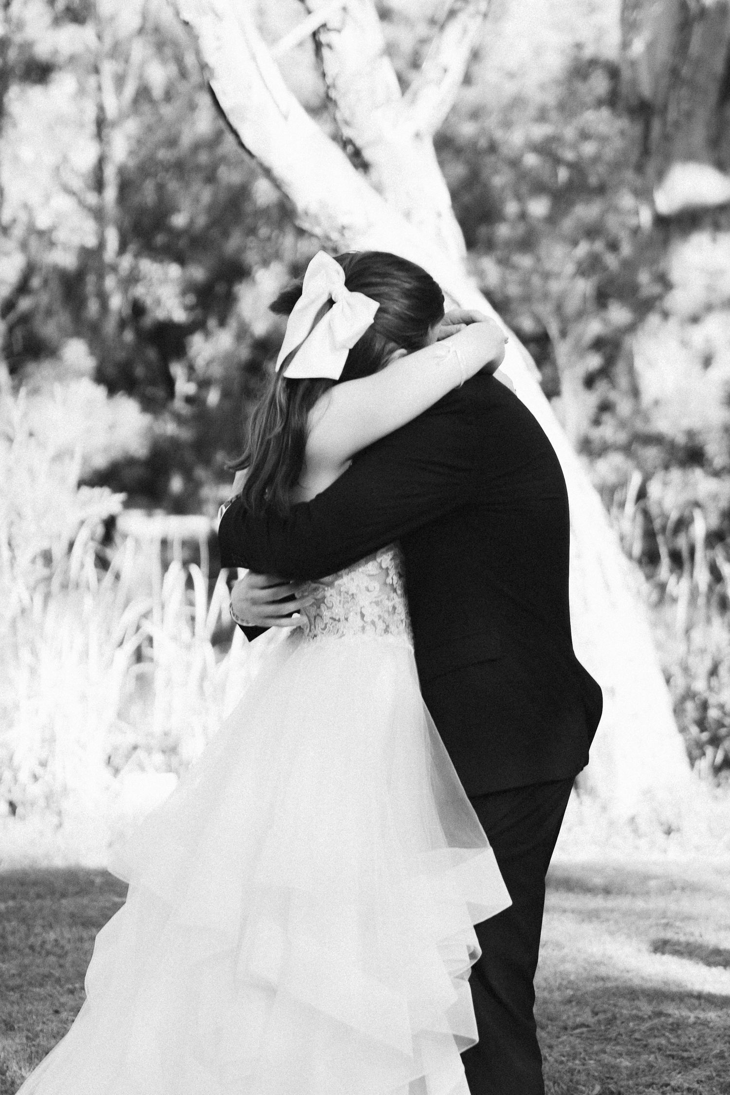 Black and white photo of bride and groom sharing a tender embrace outdoors, bride in ruffled gown with bow hair accessory.