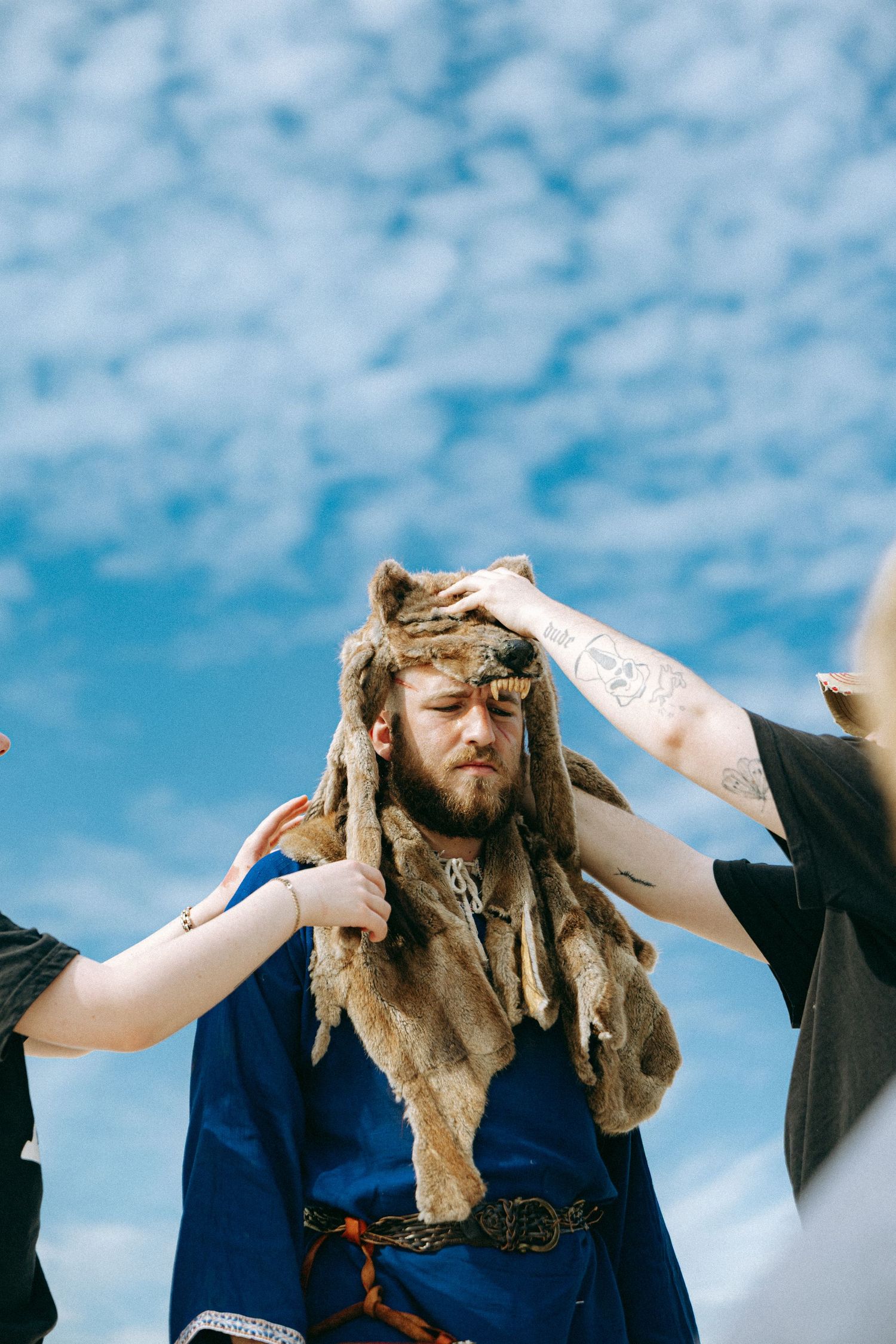 A bearded Viking warrior in blue tunic and fur bear hat being styled against a bright blue sky.