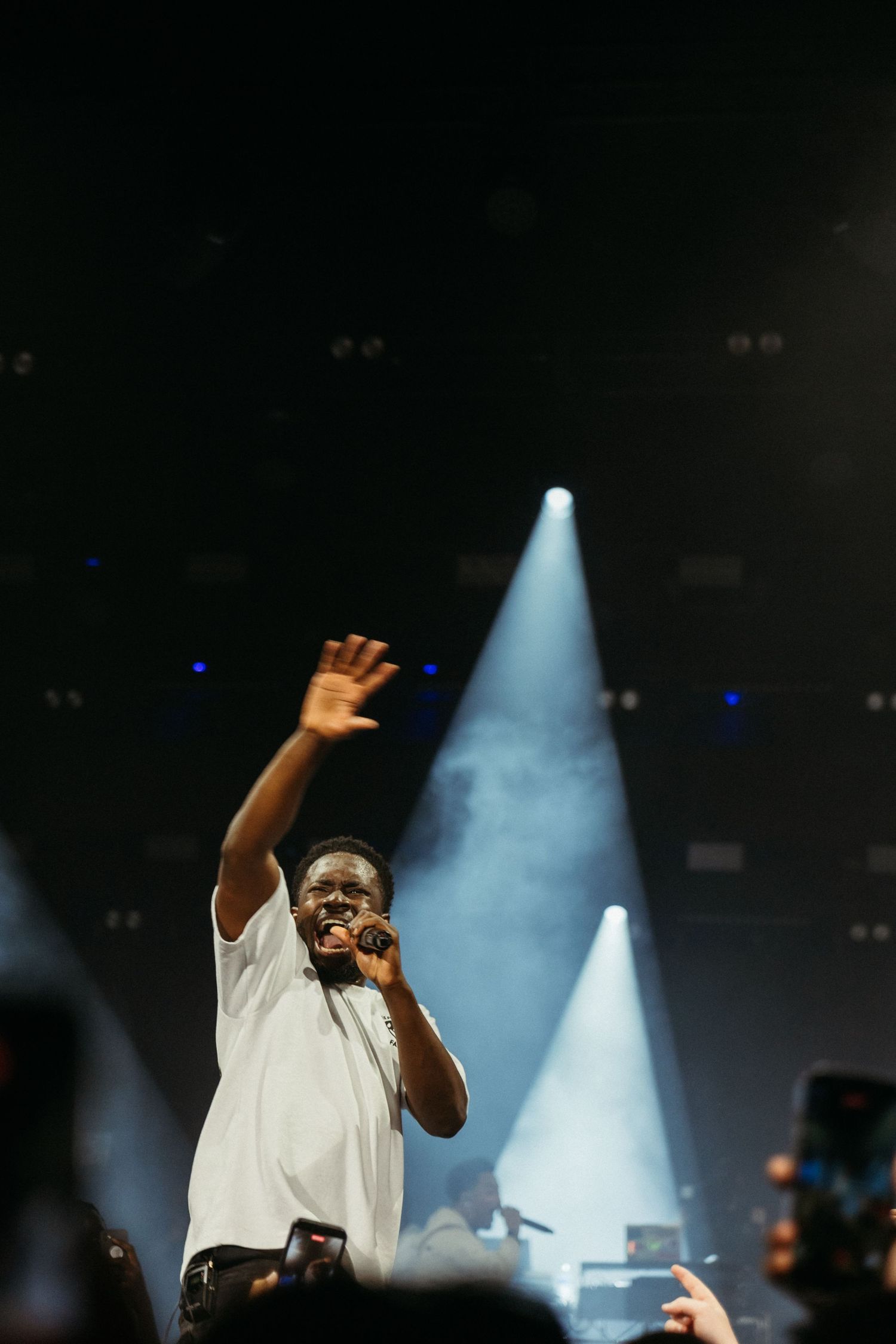 A performer in a white t-shirt sings into a microphone while raising one hand on a dramatic concert stage with blue spotlights.
