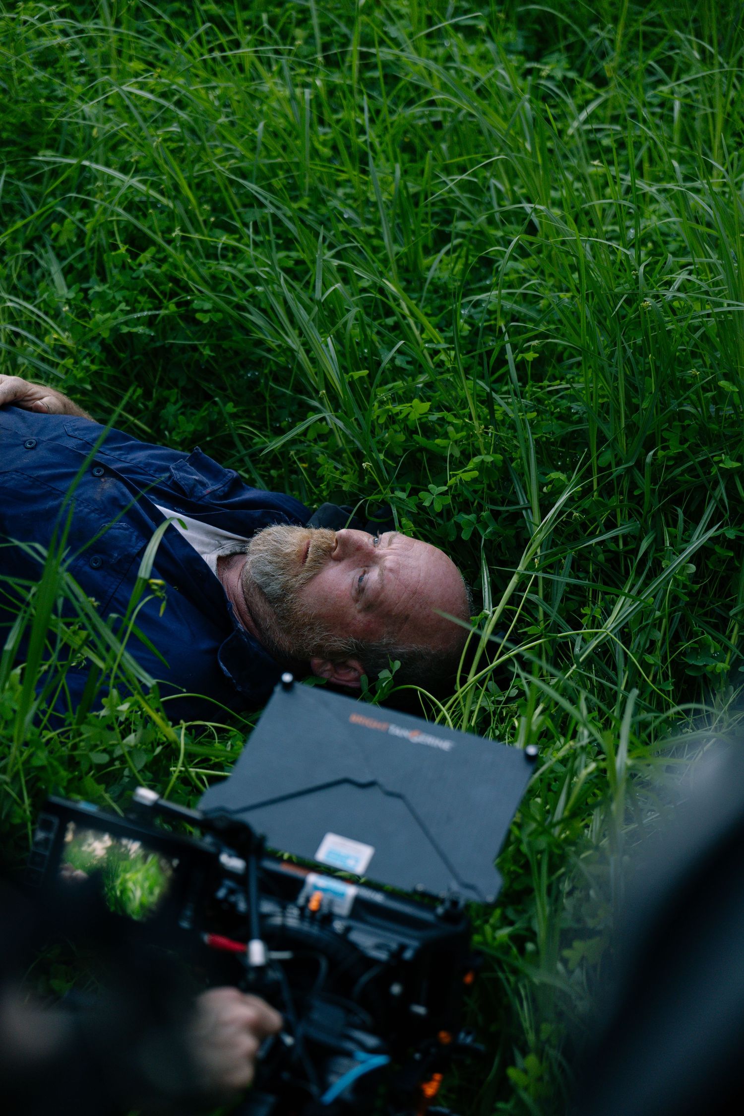 Behind-the-scenes film shoot with bearded actor lying in tall green grass while camera films.
