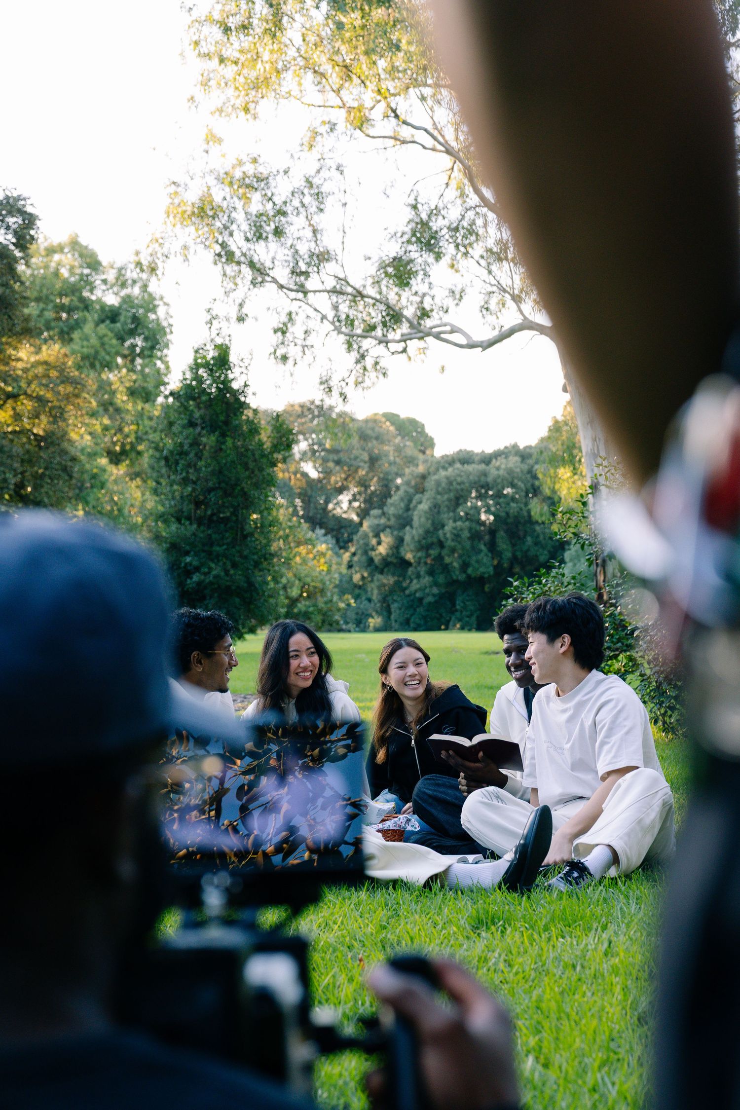 Diverse group of young people sitting on grass in a park, smiling and reading together.