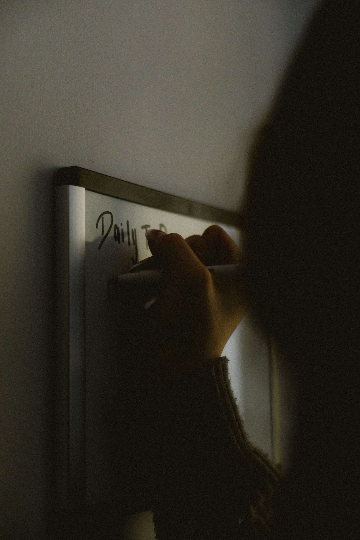 Close-up of a hand writing on a whiteboard labeled Daily Tasks in a dimly lit room.