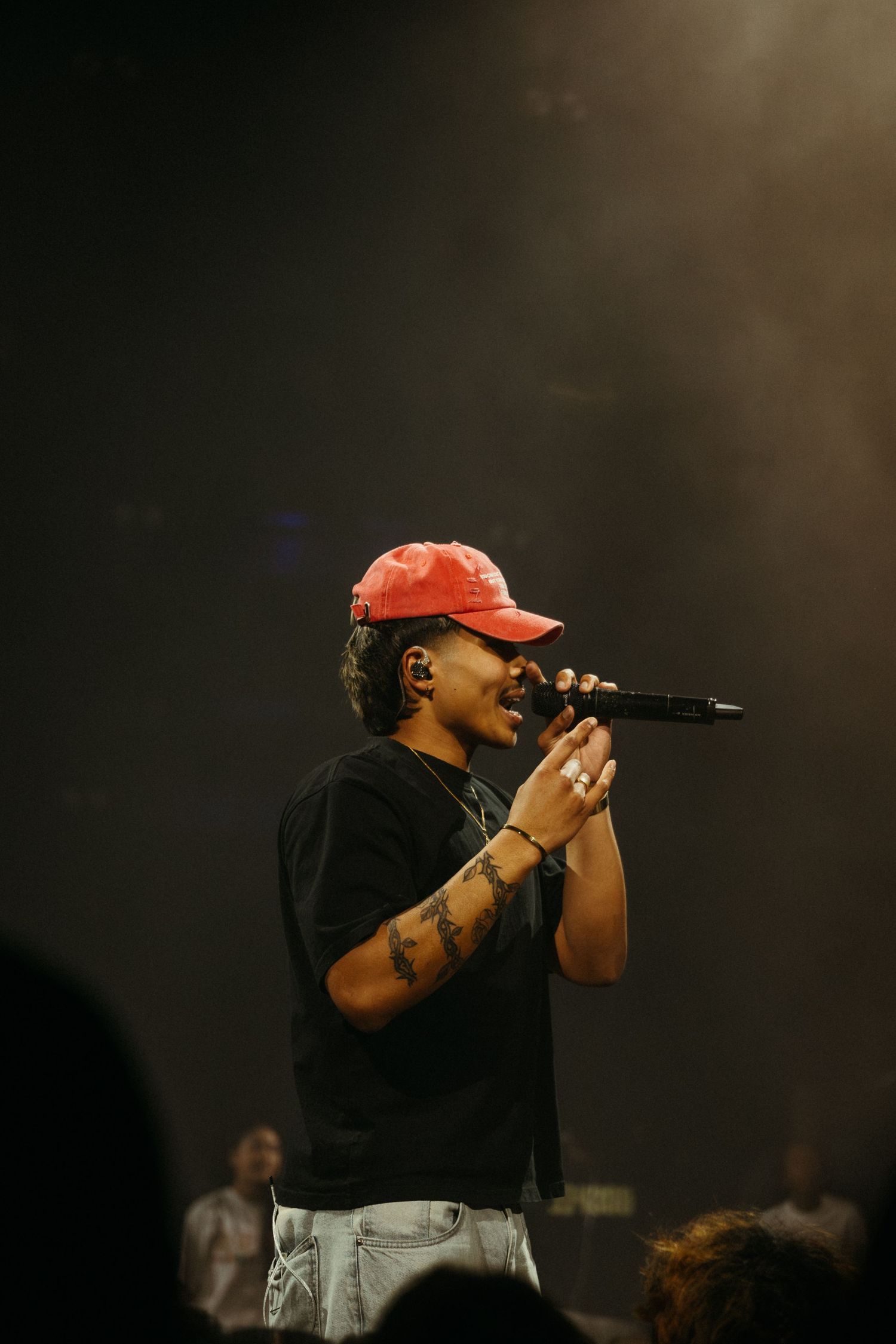 Performer in red cap and black tee sings into microphone on smoky concert stage.