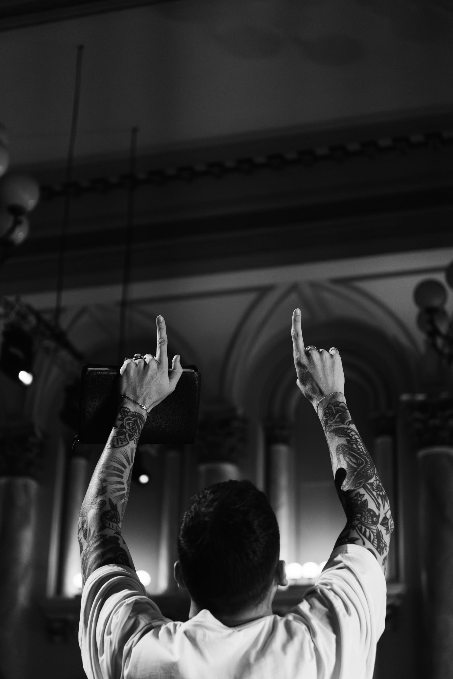 Tattooed man raising both hands pointing upward at religious worship event in church.