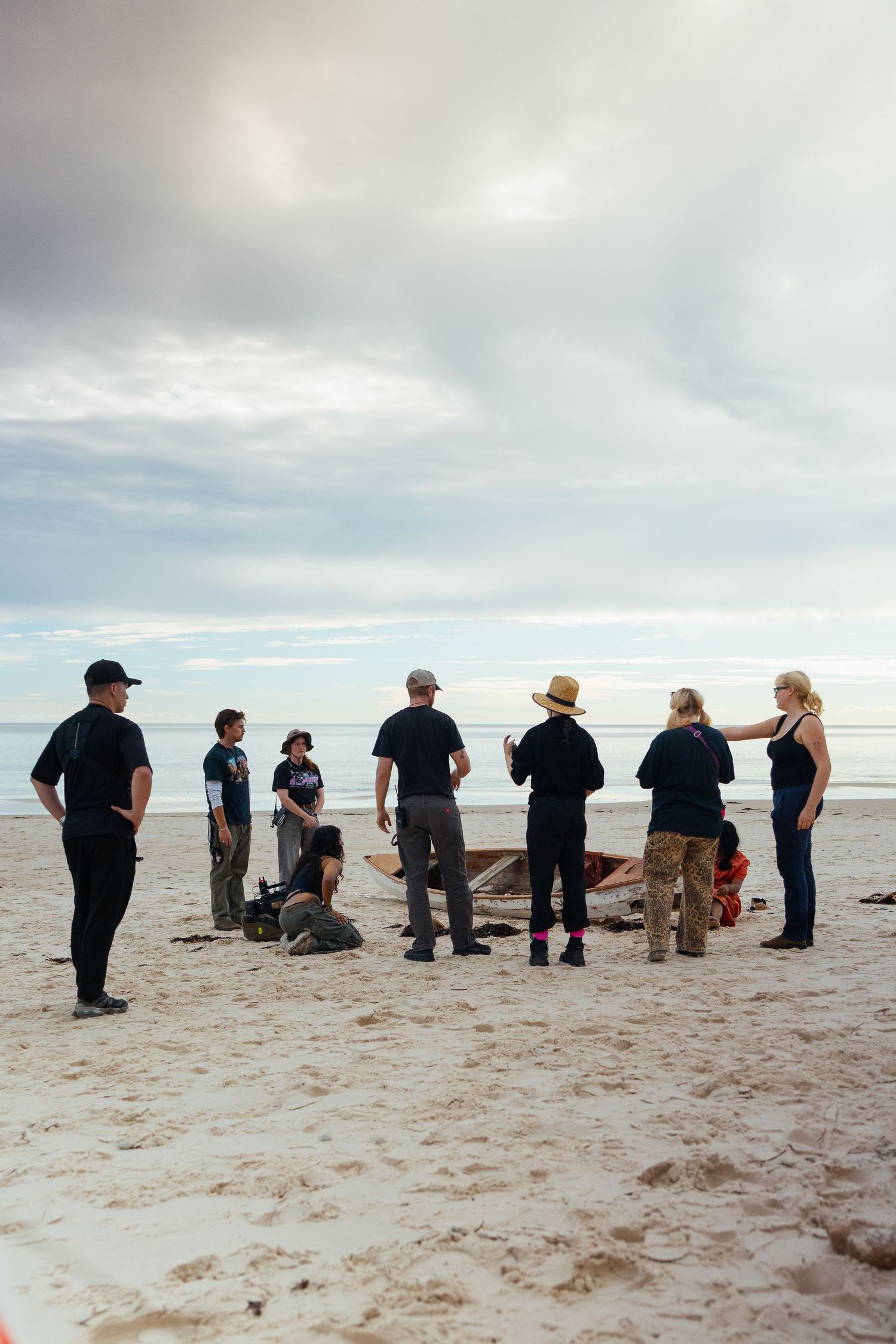 Group of people gathered on a sandy beach under a cloudy sky during a film production shoot.