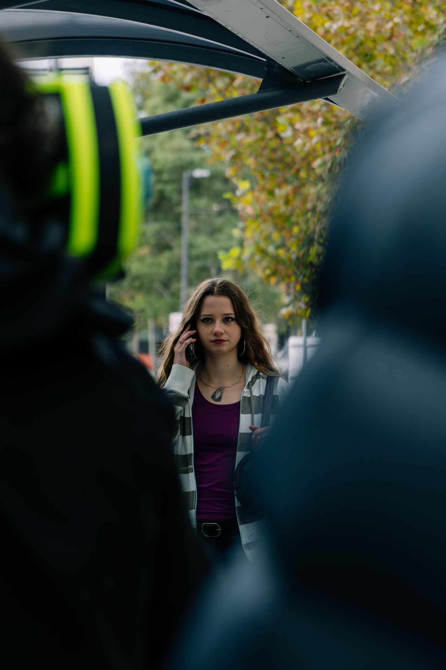 Young woman on a phone call outdoors framed between blurred foreground figures near a vehicle.