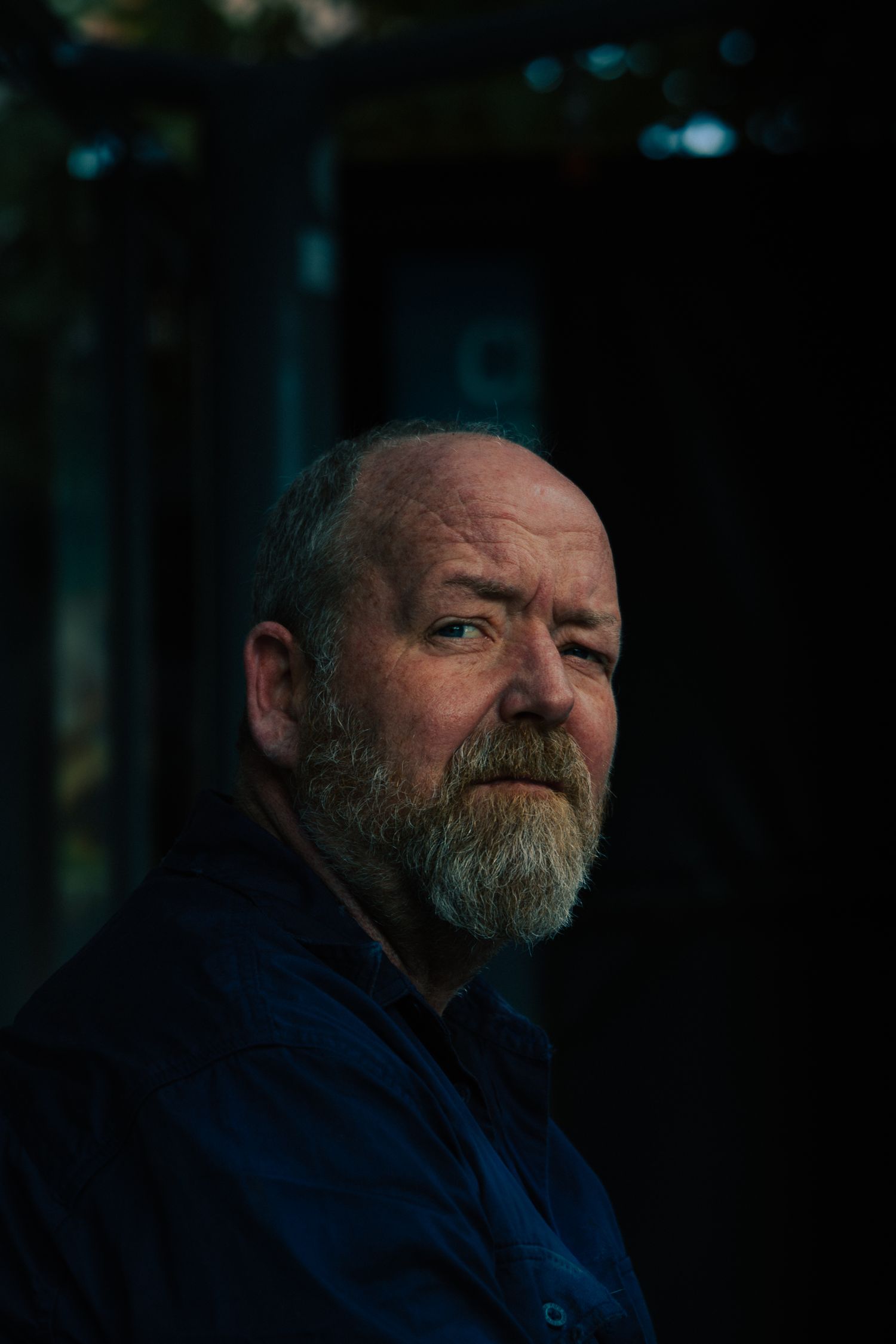 Moody portrait of a middle-aged bearded man with grey hair against a dark background.