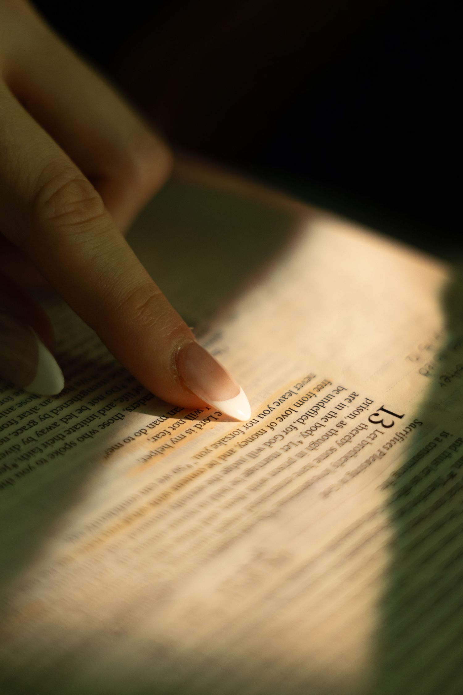 A woman's finger with manicured nails pointing to highlighted text in a book illuminated by warm light.