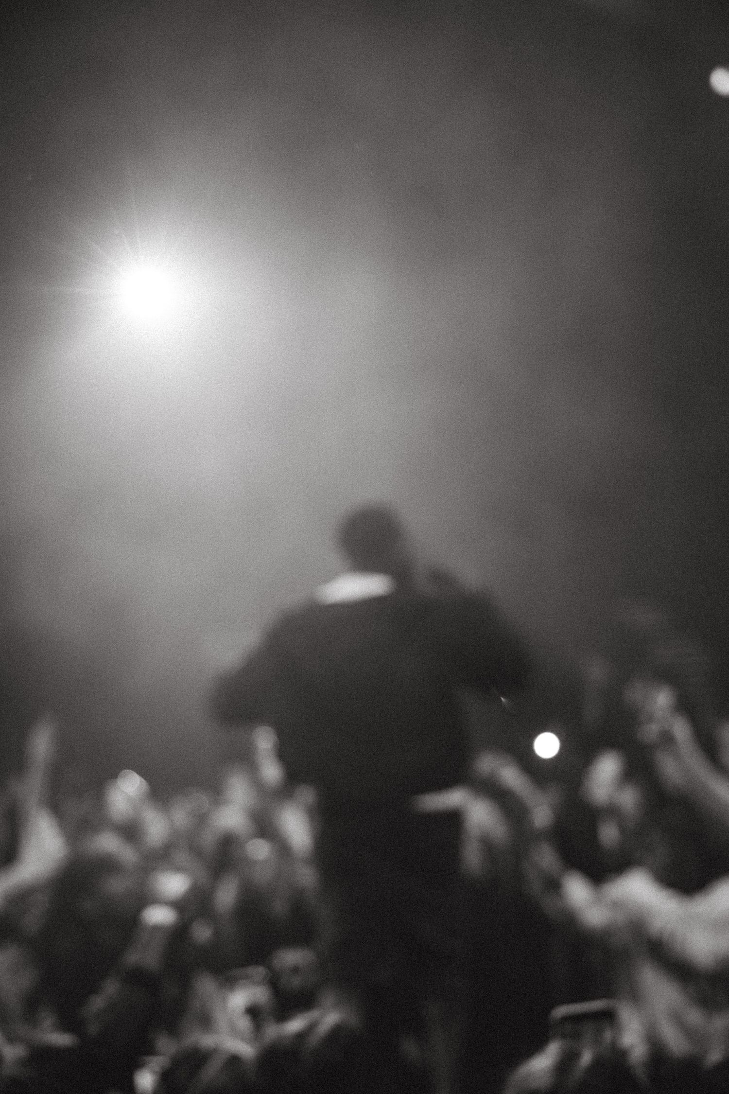 Blurry black and white concert photo of a performer on stage with raised hands crowd and bright spotlight.