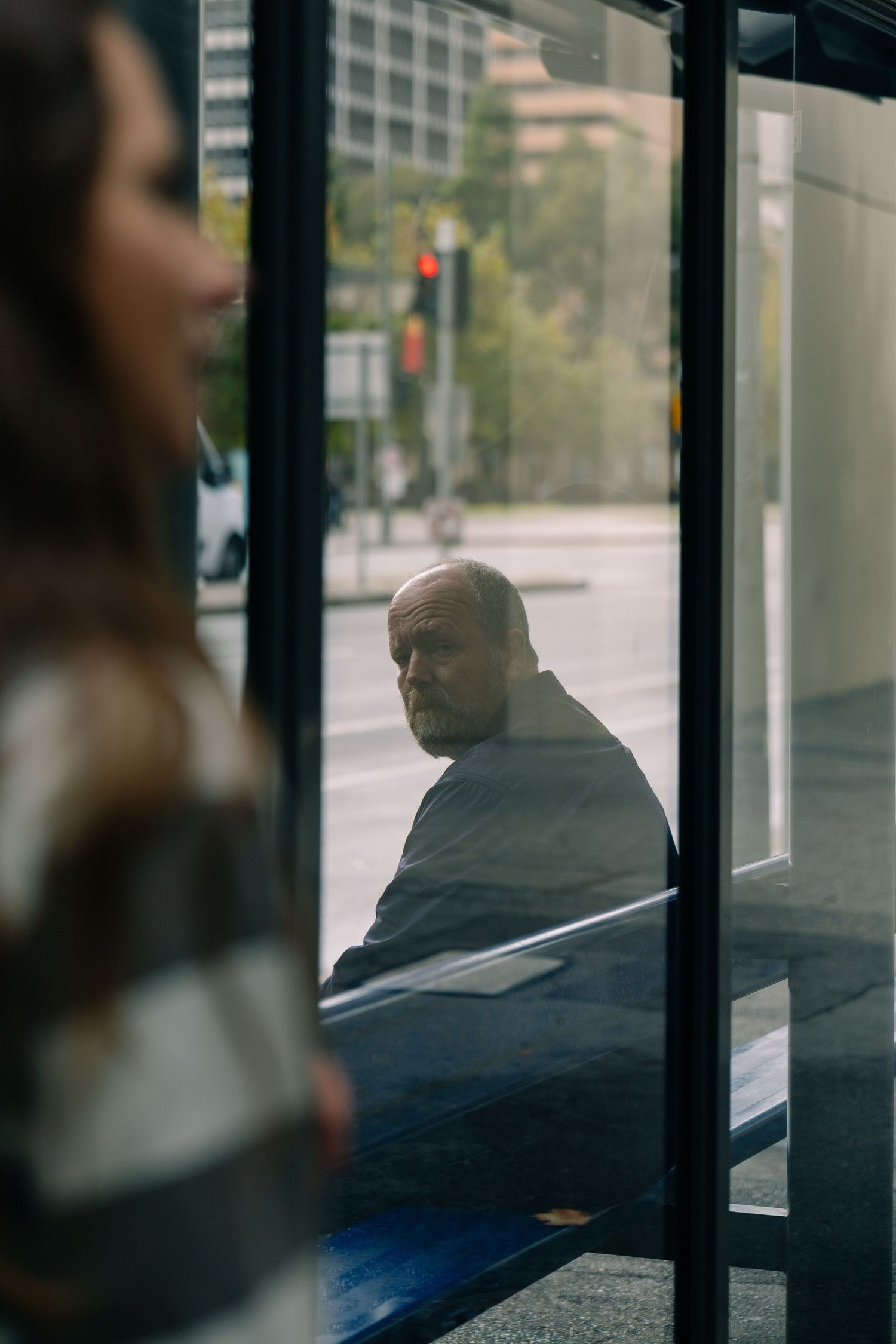 Bald man seen through glass door reflection at urban bus stop with city street behind.