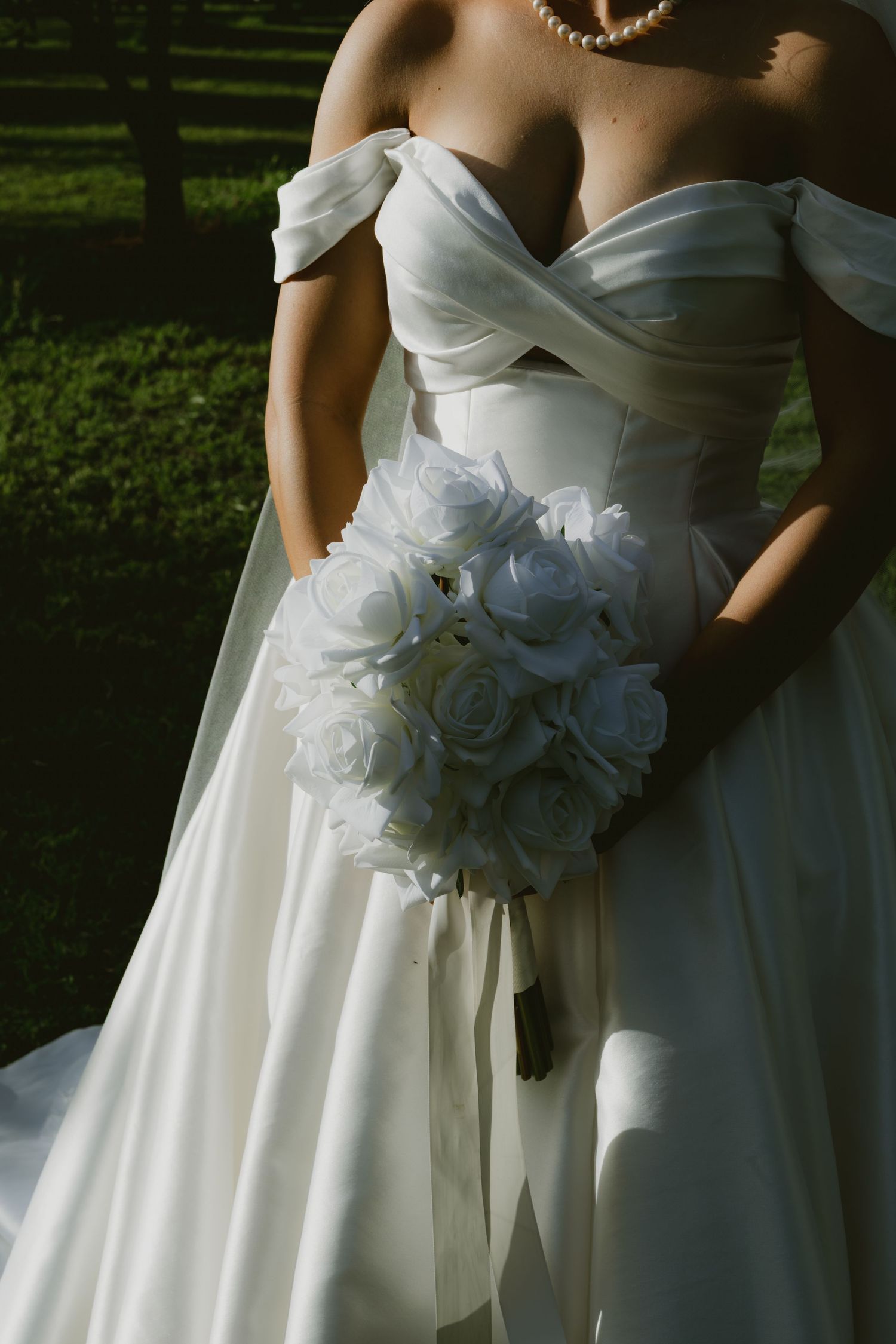 Bride in white off-shoulder gown holding elegant white rose bouquet with ribbon, standing outdoors in sunlight.